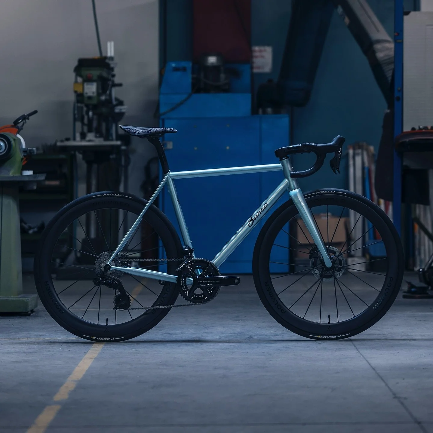 A sleek silver road bike with black wheels and tires, set inside a workshop or garage with tools and equipment in the background.