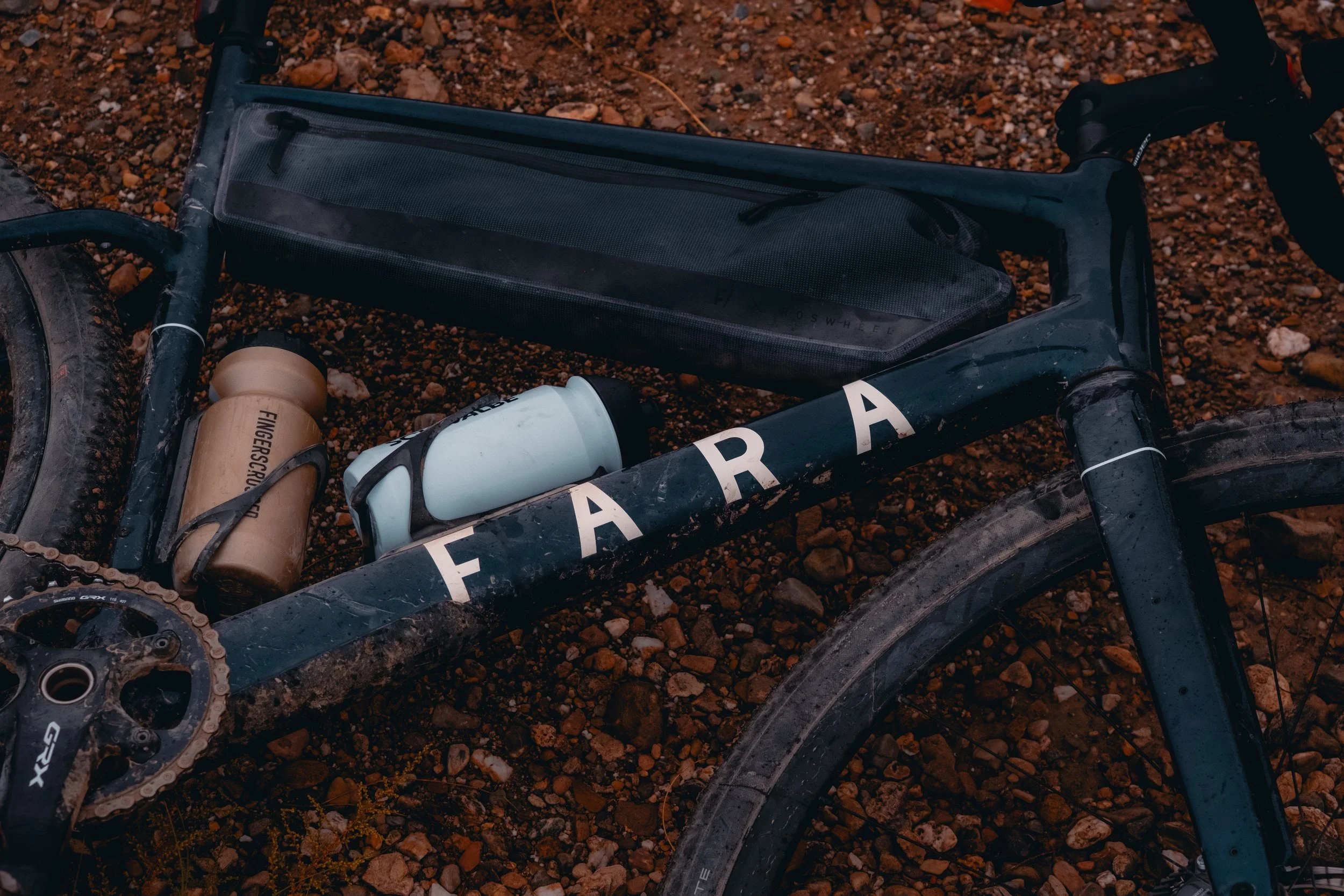 A black Fatbike with dirt on the frame, lying on rocky ground. The bike has two water bottles, one brown and one light blue, attached to the frame.
