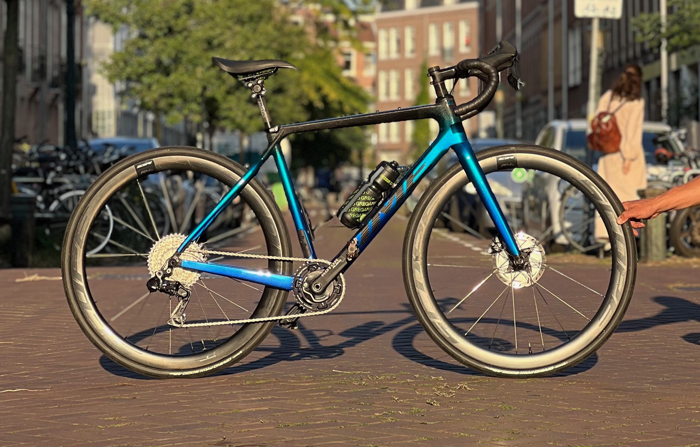 A blue and black mountain bike on a red paved street, with a row of parked bicycles and buildings in the background, and an arm reaching towards the rear wheel on the right side.