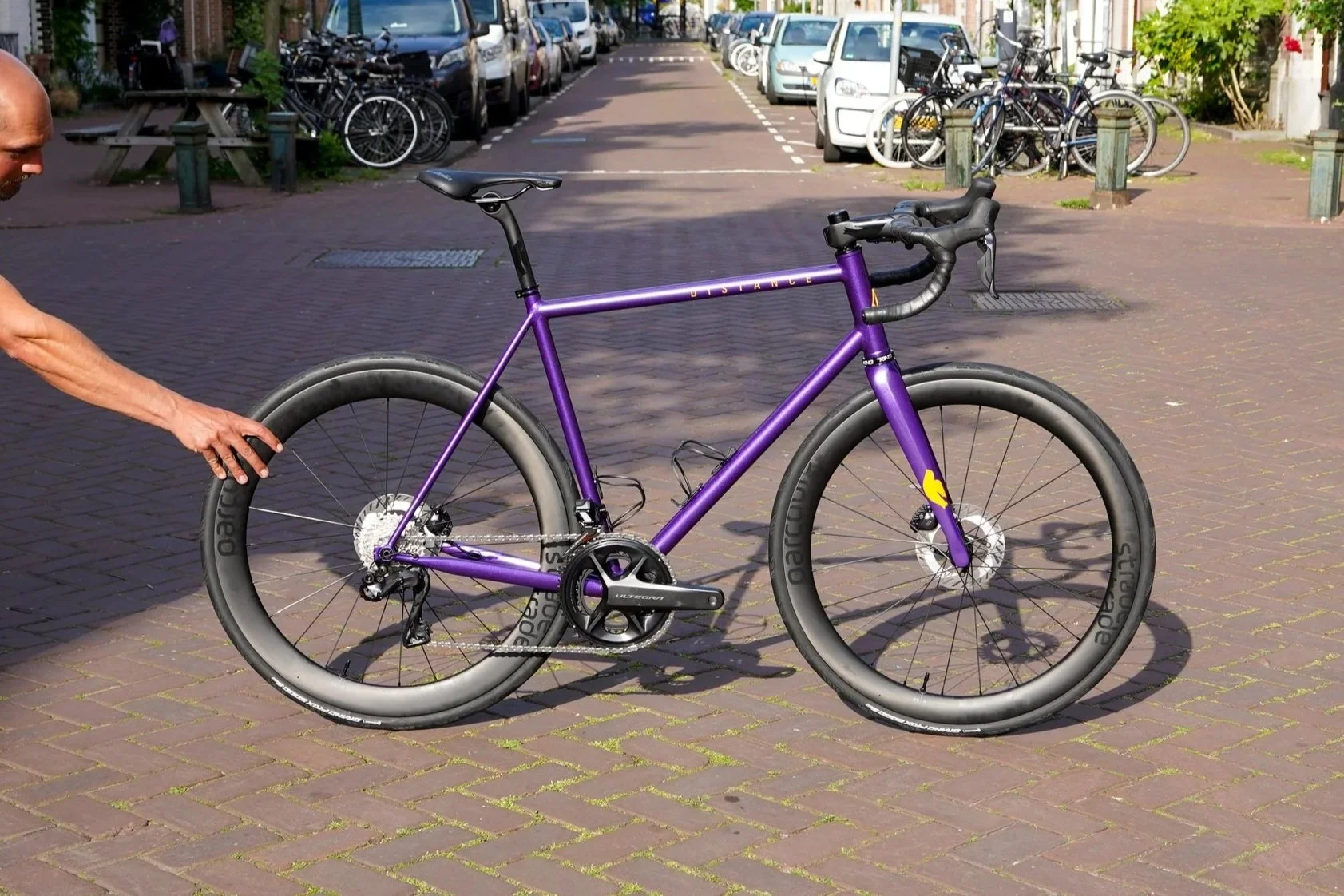 Person holding a purple gravel bike with thick tires on a brick-paved street, parked alongside other bikes and cars in the background.