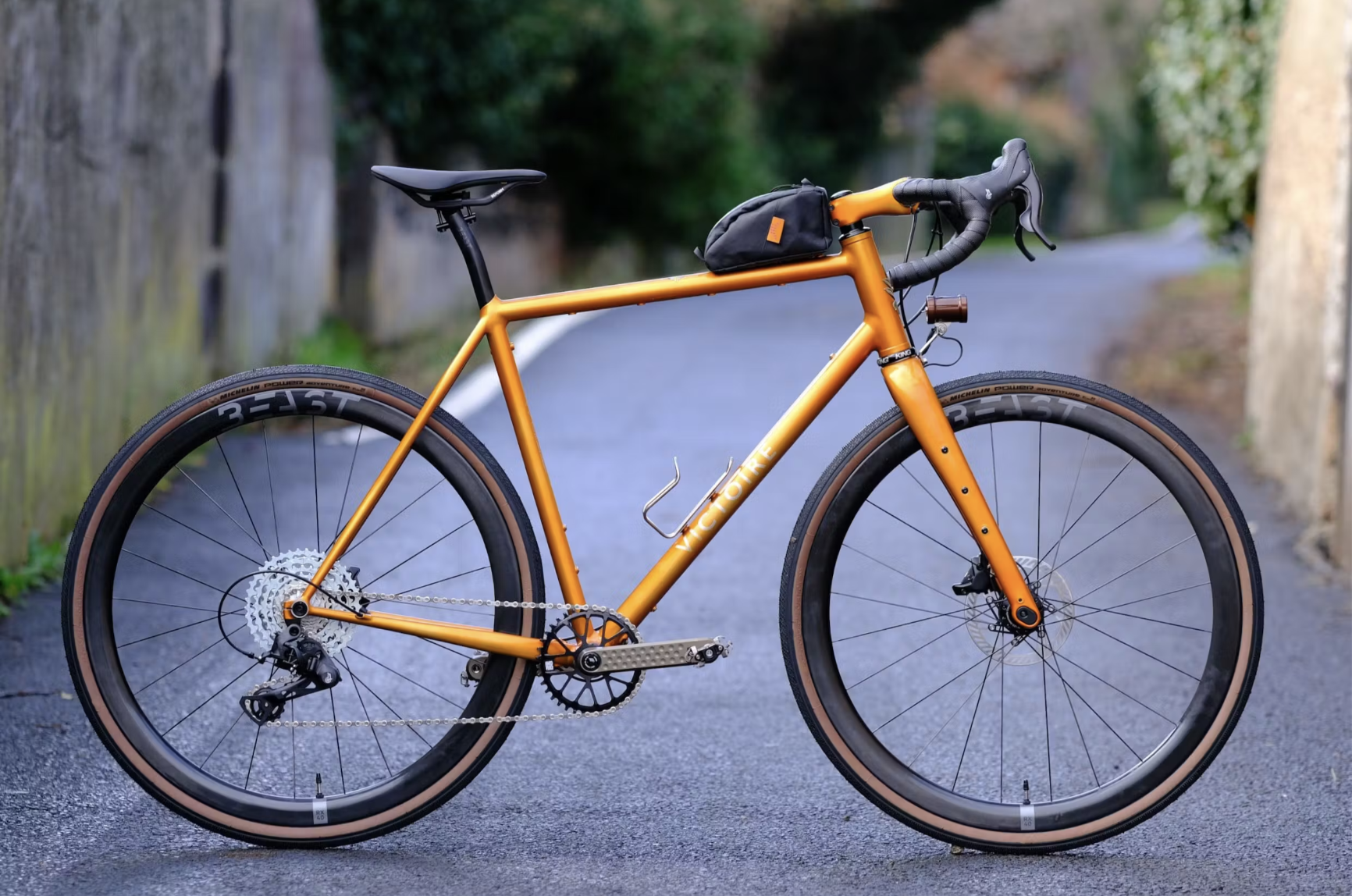 An orange mountain bike with black tires and a small black bag attached to the handlebars, parked on a paved road.