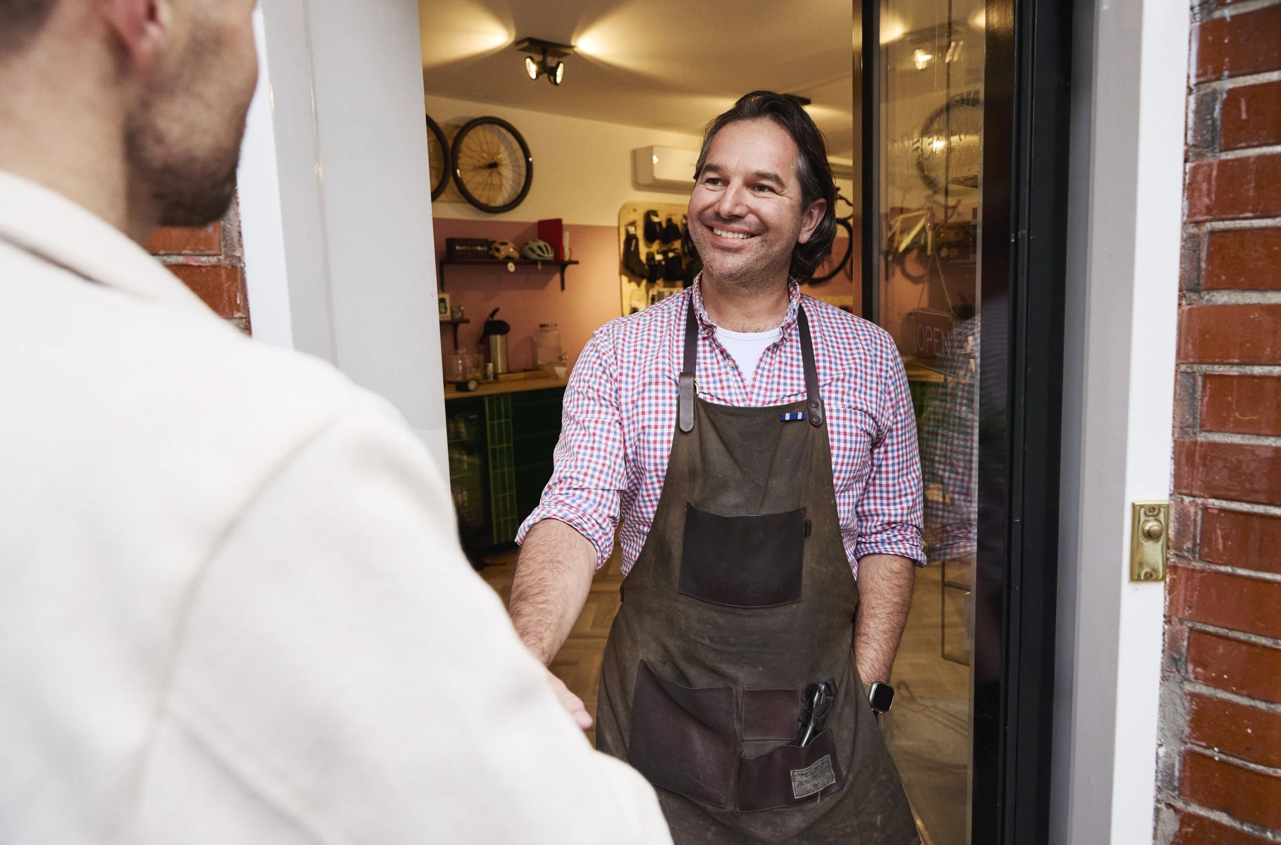 A smiling man in a checkered shirt and apron greets another person at the entrance of a cafe or shop.