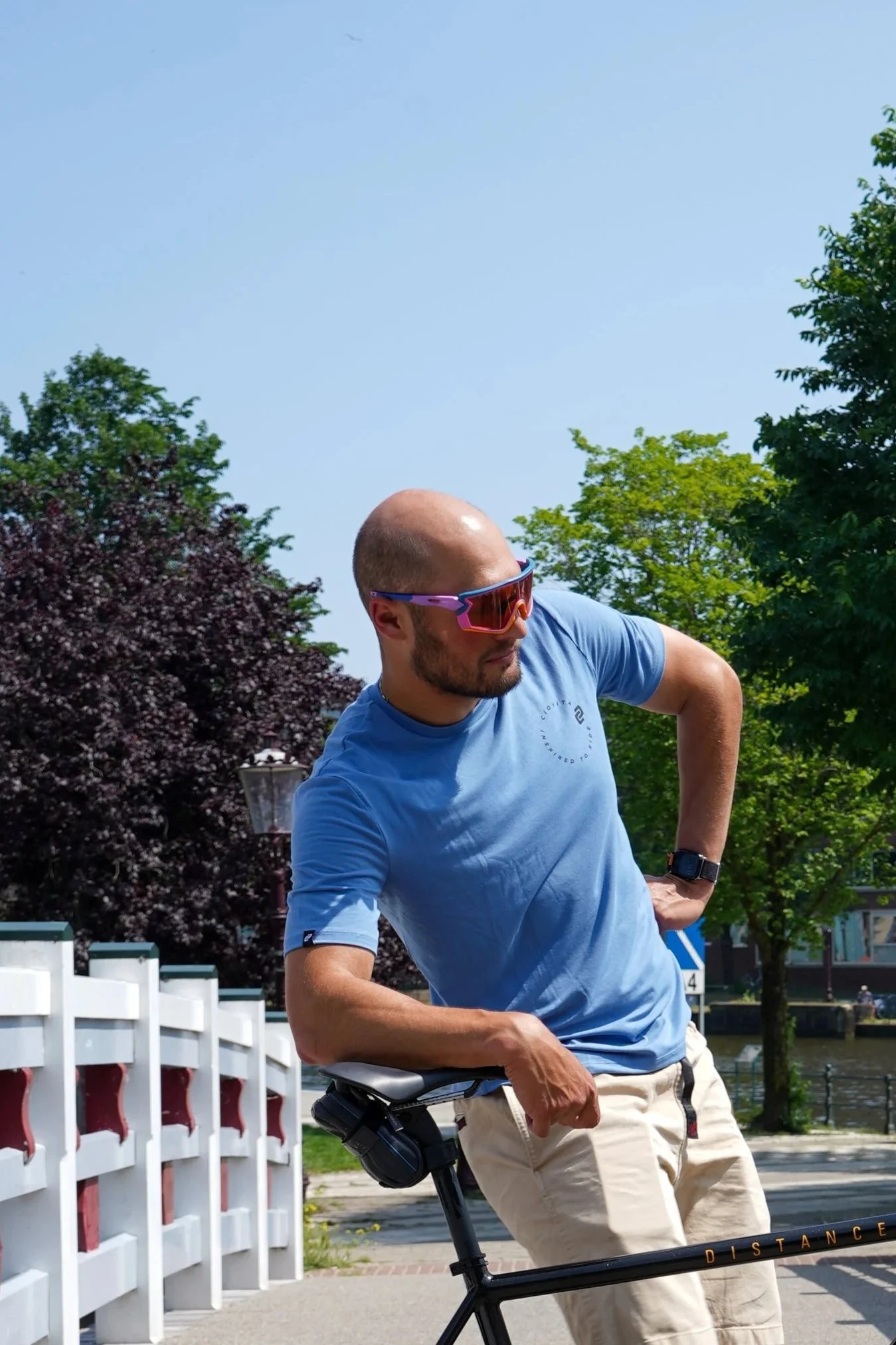 A man with a shaved head wearing sunglasses, a blue T-shirt, and beige pants leaning on a bike outdoors on a sunny day with trees and a canal in the background.