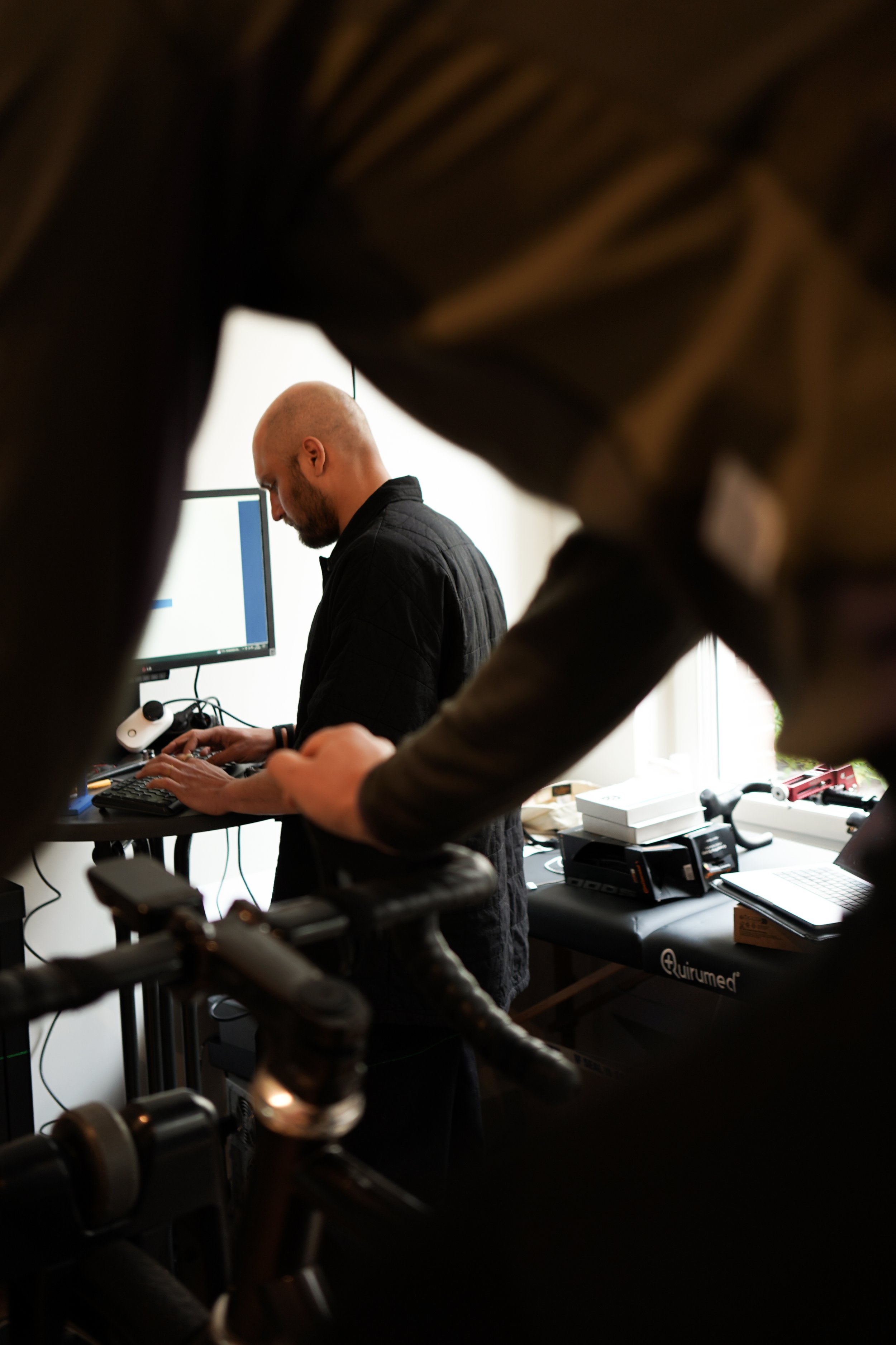 A man with a shaved head and beard working on a computer in an indoor setting, viewed through the frame of another person's legs and bicycle handlebars.