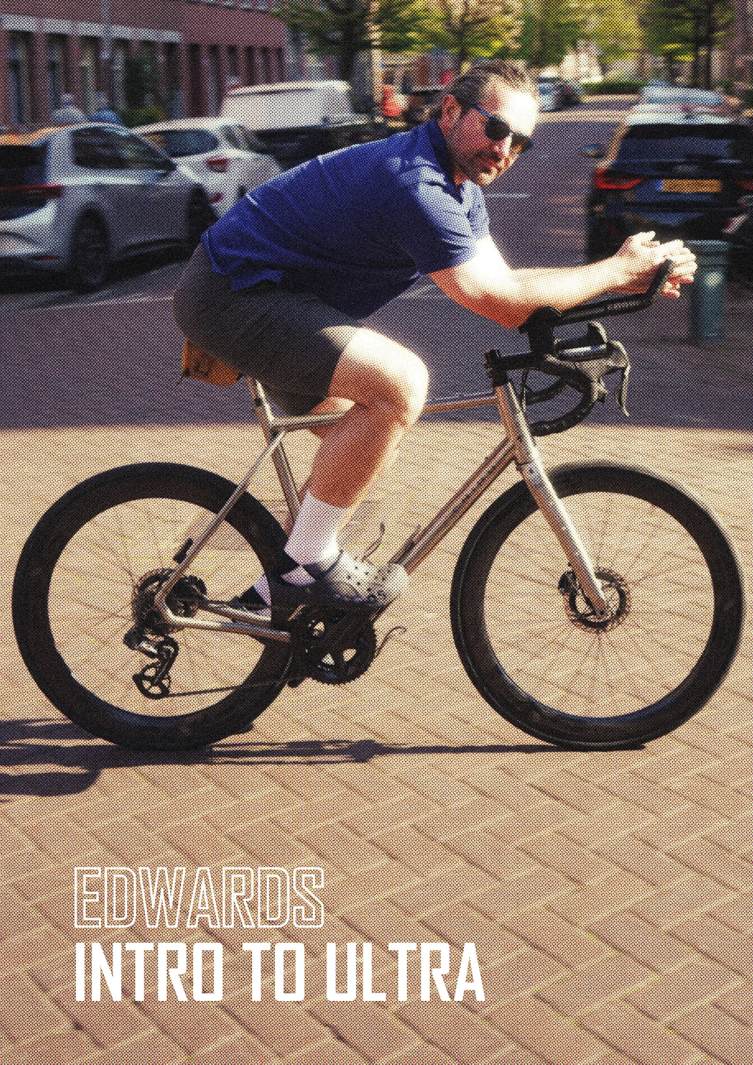 A man riding a bicycle on a brick pavement in a parking lot, wearing sunglasses, a blue shirt, shorts, and white socks. There are parked cars and buildings in the background.