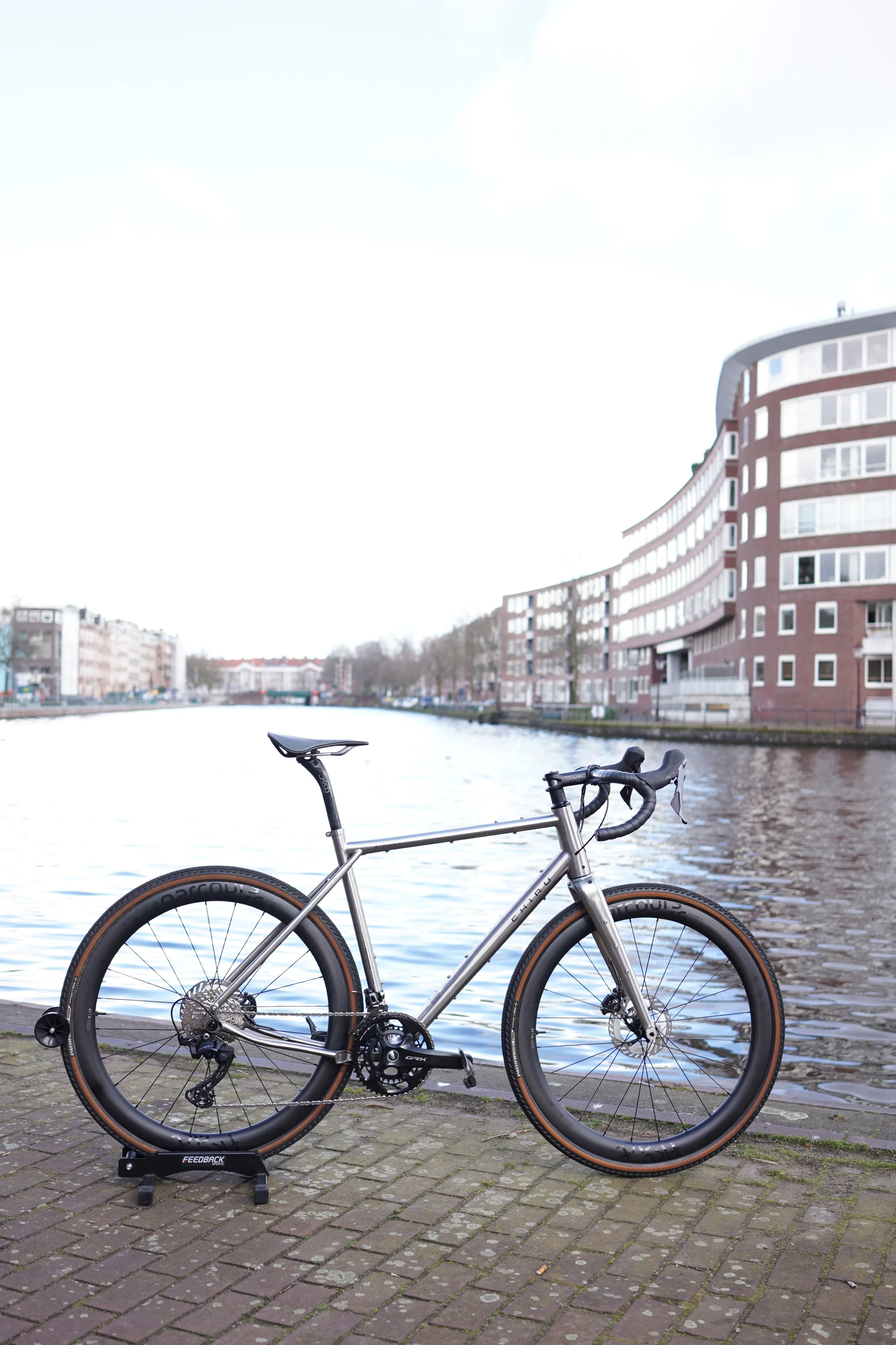 A bike parked on a paved path by a river with modern apartment buildings in the background on a cloudy day.