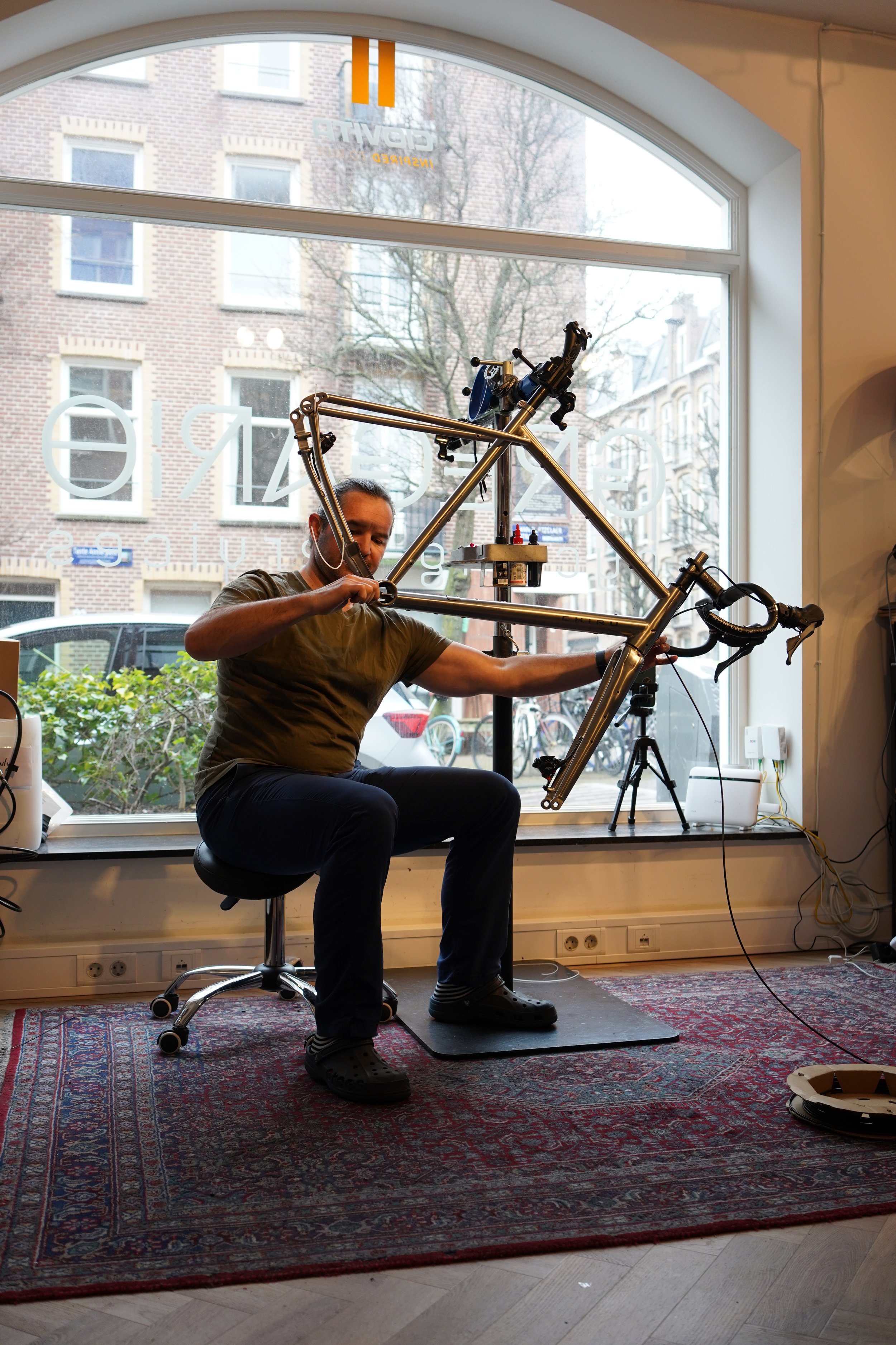 A man working on a bicycle inside a shop, sitting on a stool in front of a large window, with tools and equipment around him.