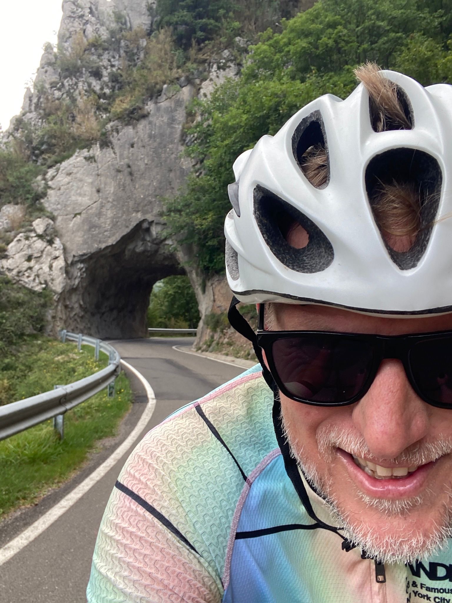 Close-up of a smiling man wearing a white bicycle helmet and black sunglasses, riding a bike on a mountain road with a tunnel in the rocky hillside in the background.