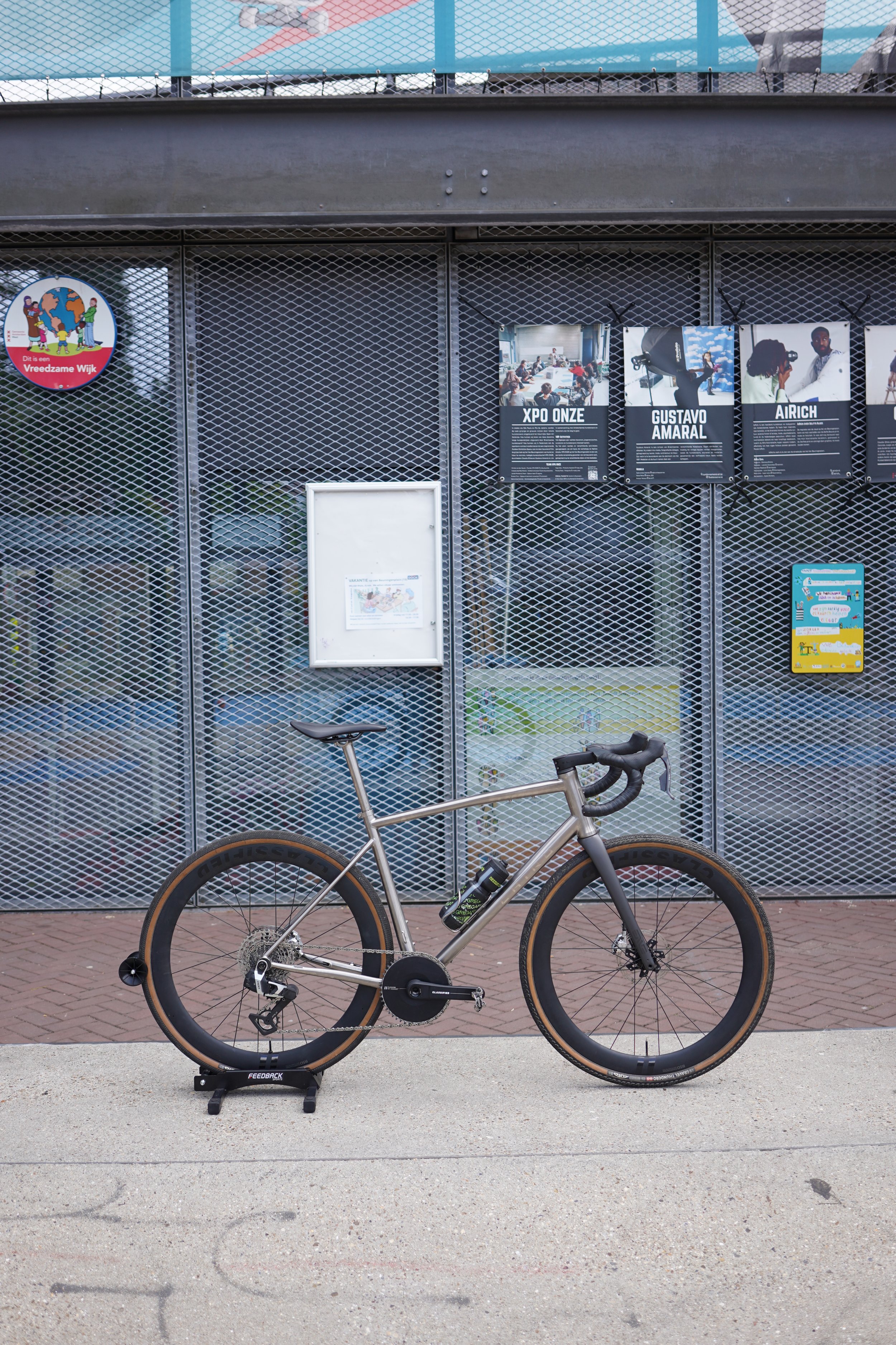 A bicycle parked on a sidewalk in front of a metal fence with posters and signs. One sign has a colorful logo with children holding hands.