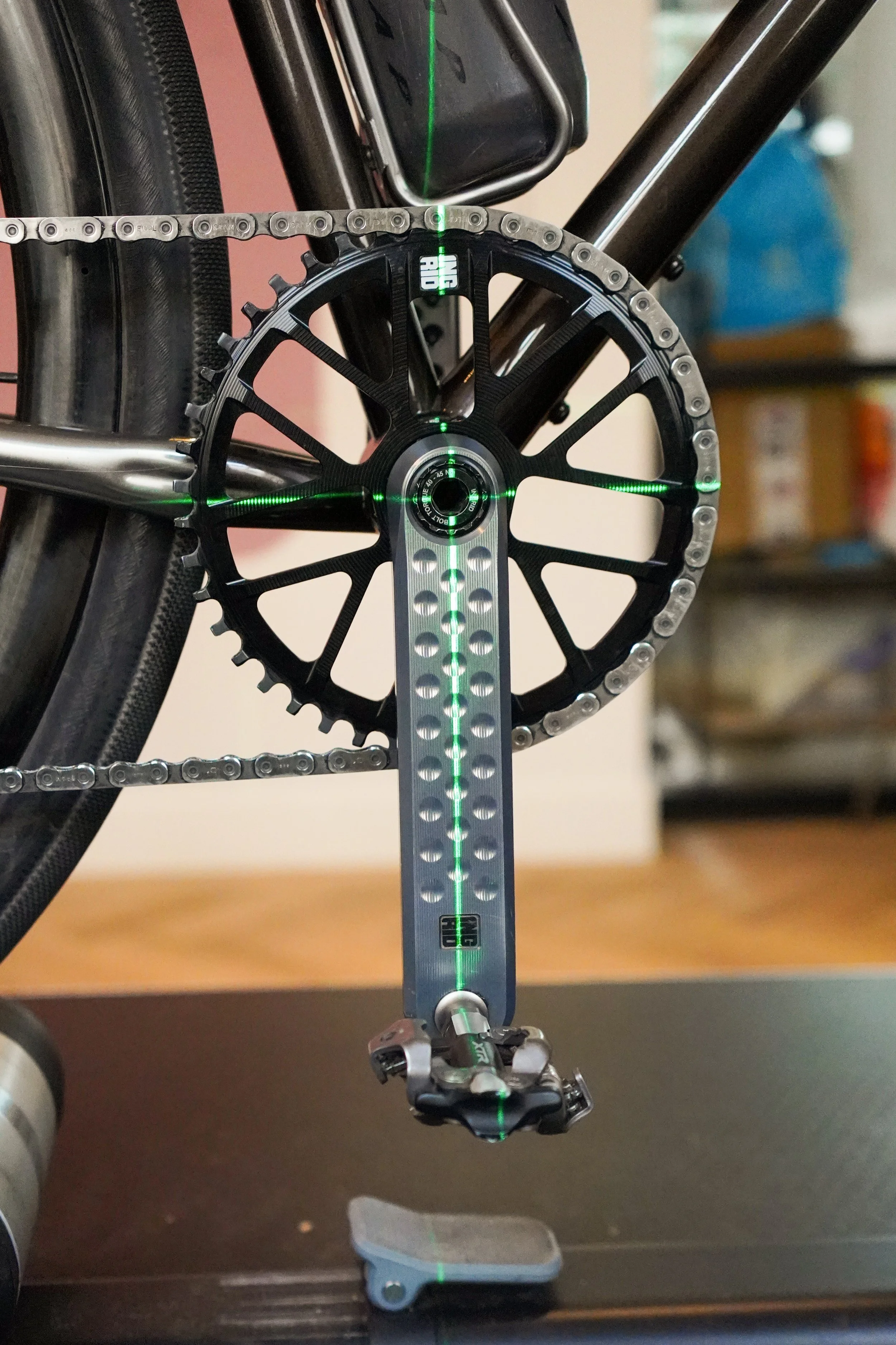 Close-up of a bicycle's crankset and chain with laser alignment dots projected on the chainring, crank arm, and rear wheel.