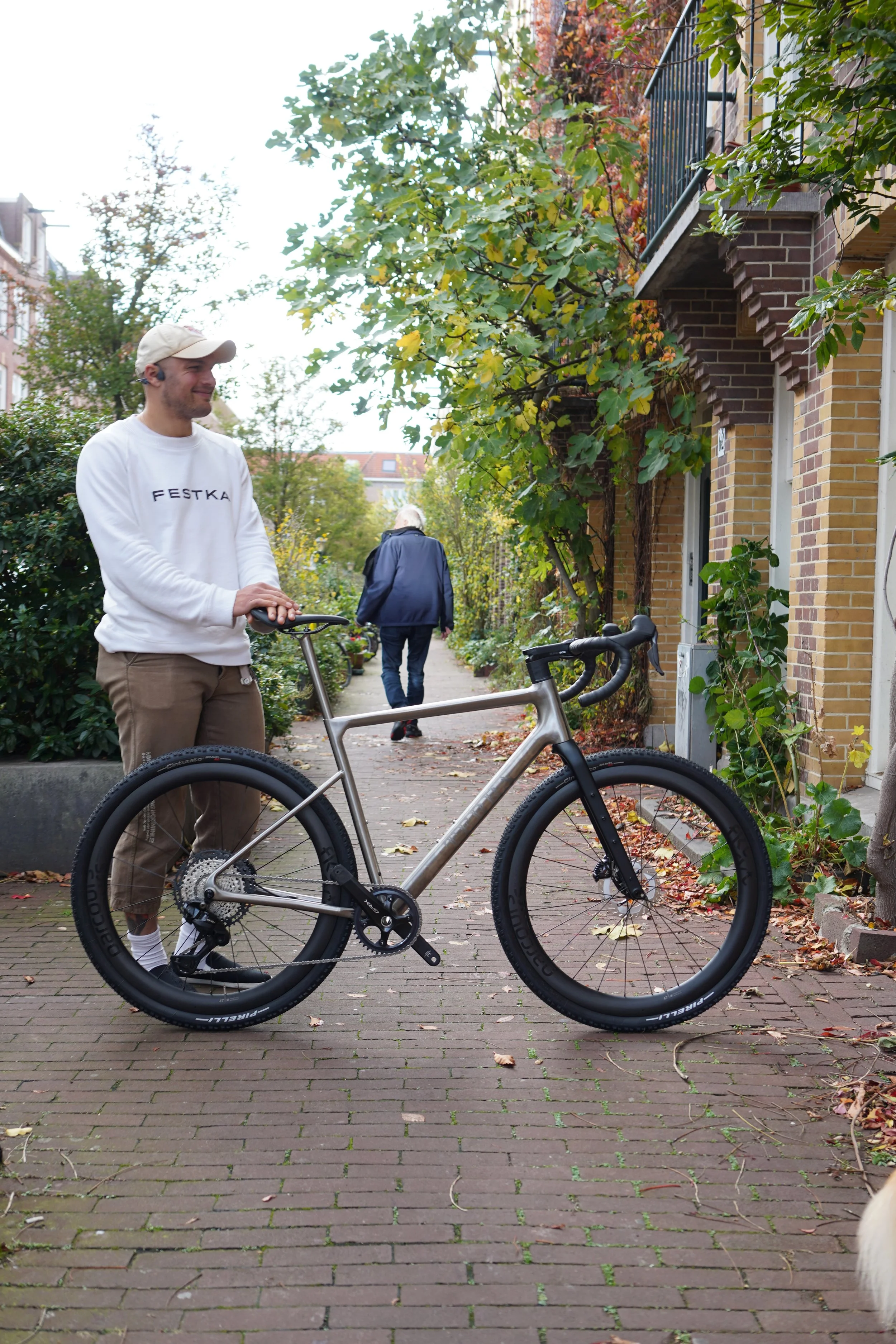A smiling young man in a white sweatshirt and beige cap standing next to a modern bicycle on a brick sidewalk in a residential neighborhood with trees and shrubs.