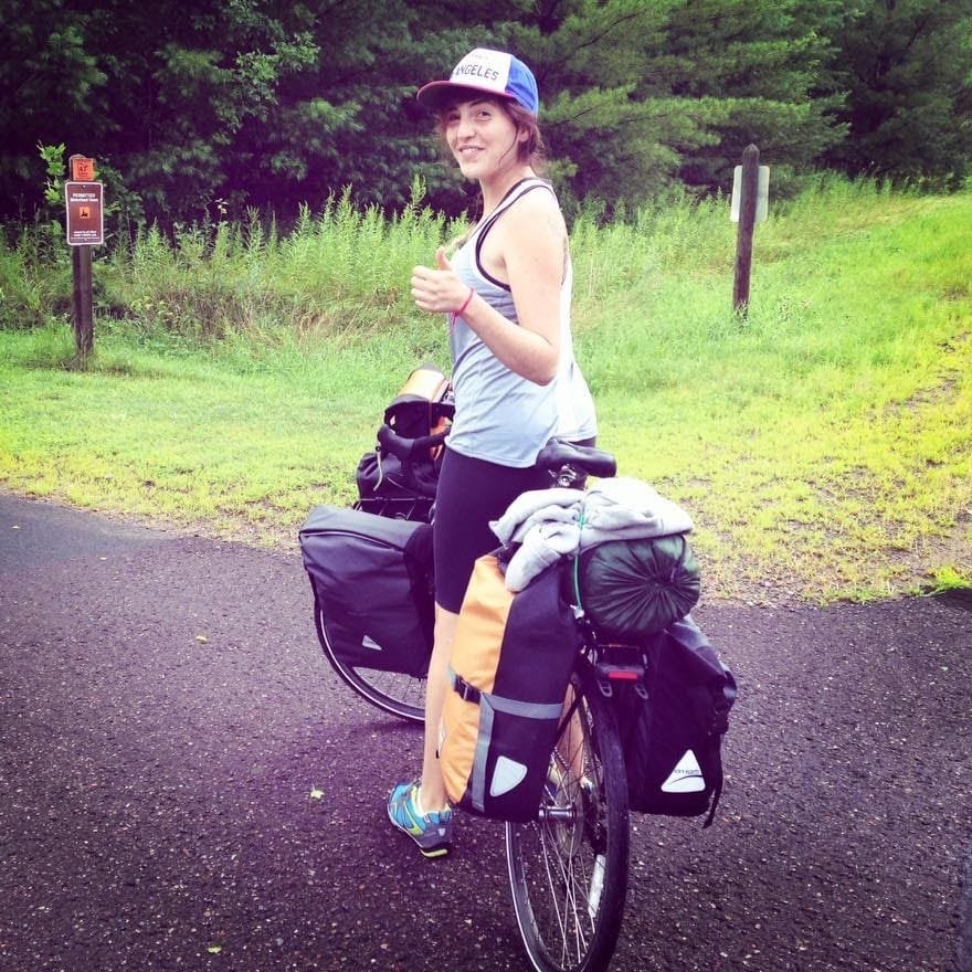 Young woman with a backpack and camping gear on a bicycle, standing on a paved trail in a park with green trees and grass.