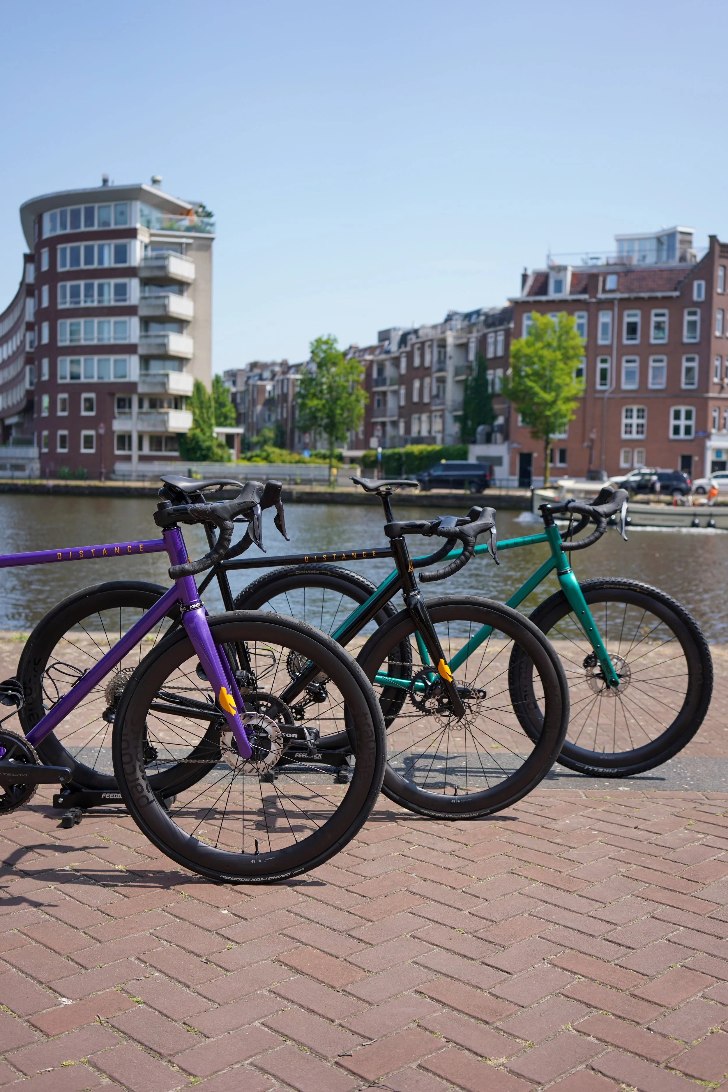 Two bicycles parked on a brick pathway by a canal, with residential buildings and trees in the background on a sunny day.