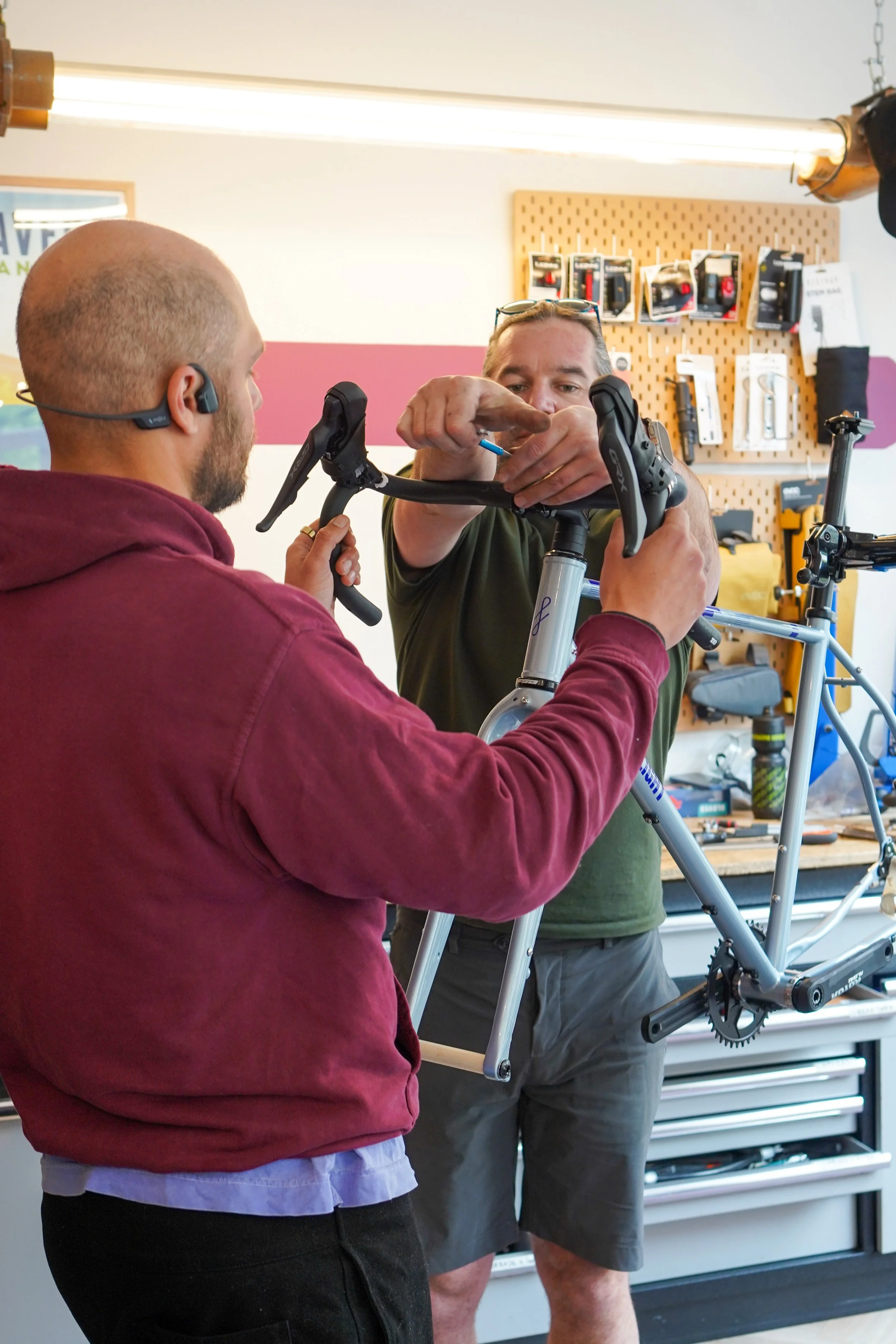 Two men working on assembling a bicycle in a workshop. One man is holding the bike frame, while the other is using a tool on the handlebars.