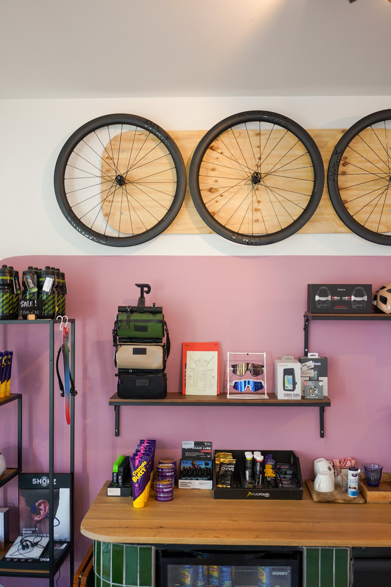 Store display with bike wheels on the wall, shelves with supplements, sunglasses, smartwatch, and accessories, a table with horse balm and other products, and pink walls.