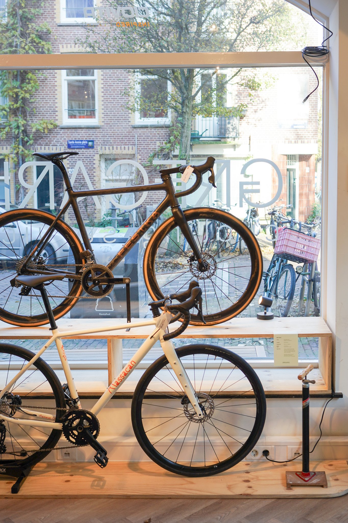 Interior of a bicycle shop with two bicycles on display, one mounted on a wooden stand and the other on a black bike stand. The window shows a street scene outside with trees, bicycles, and buildings.