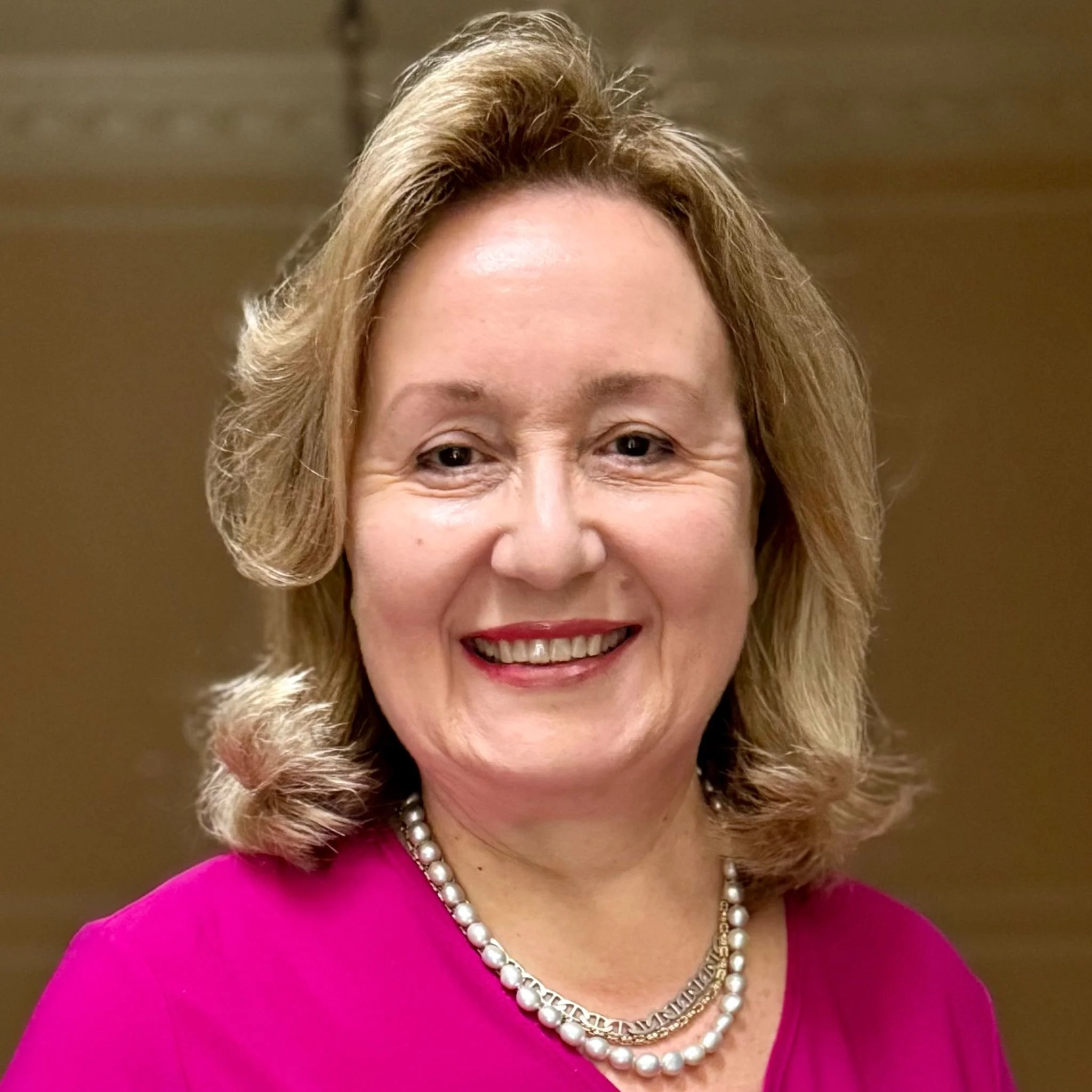 Portrait of a woman smiling, wearing a black and pink patterned top and a pearl necklace.