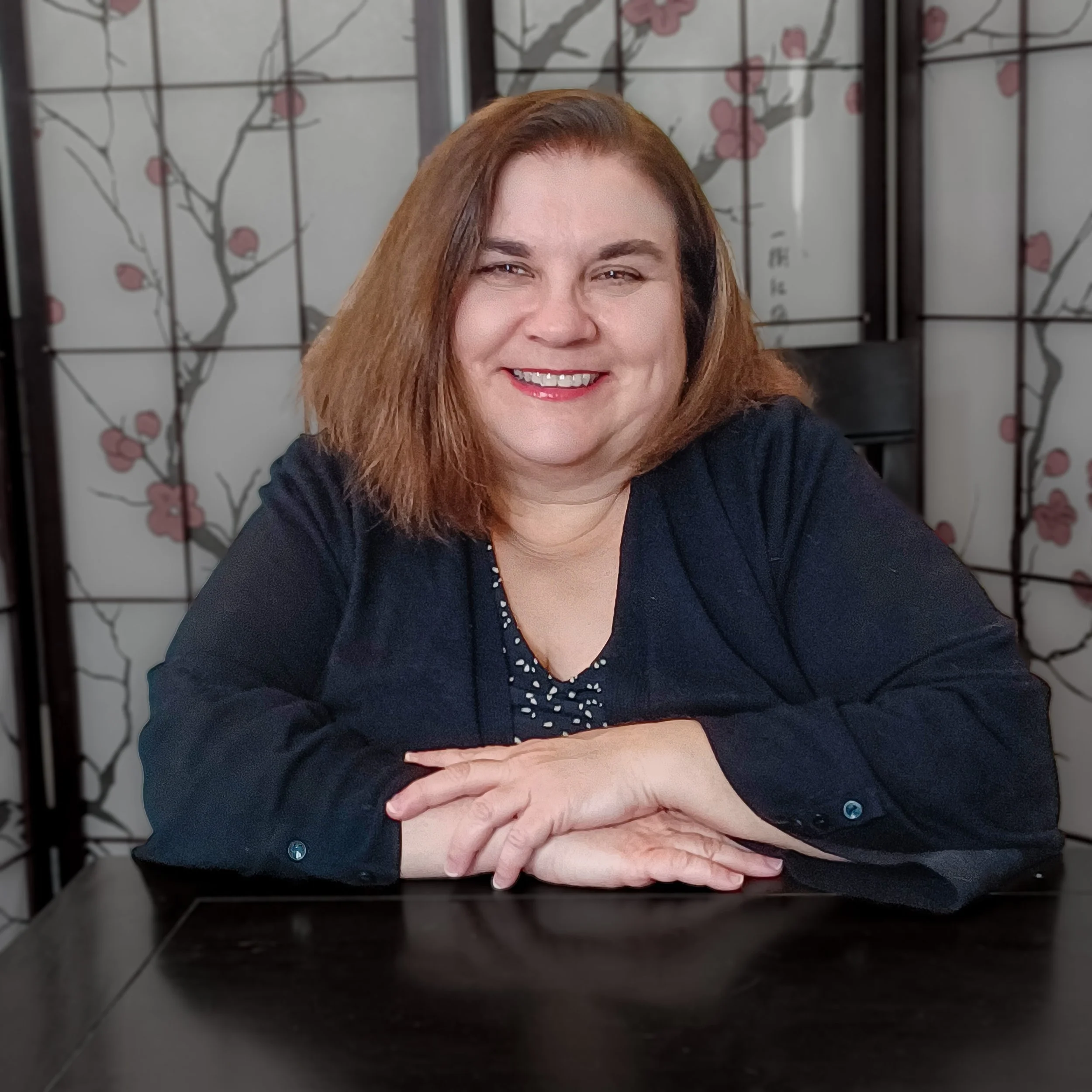 A woman with shoulder-length brown hair smiling, wearing a gray patterned top over a black shirt, against a plain background.
