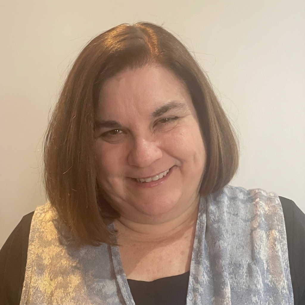 A woman with shoulder-length brown hair smiling, wearing a gray patterned top over a black shirt, against a plain background.