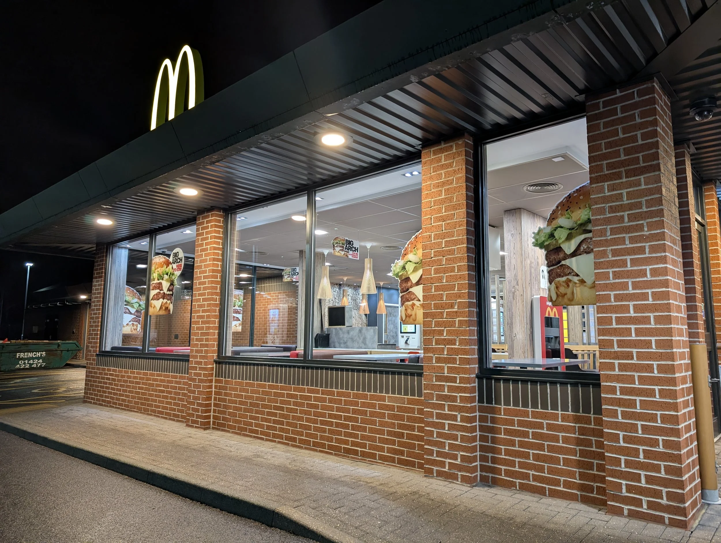 Nighttime view of a McDonald's fast food restaurant with illuminated yellow McDonald's logo, brick exterior, large glass windows, and posters advertising burgers inside.