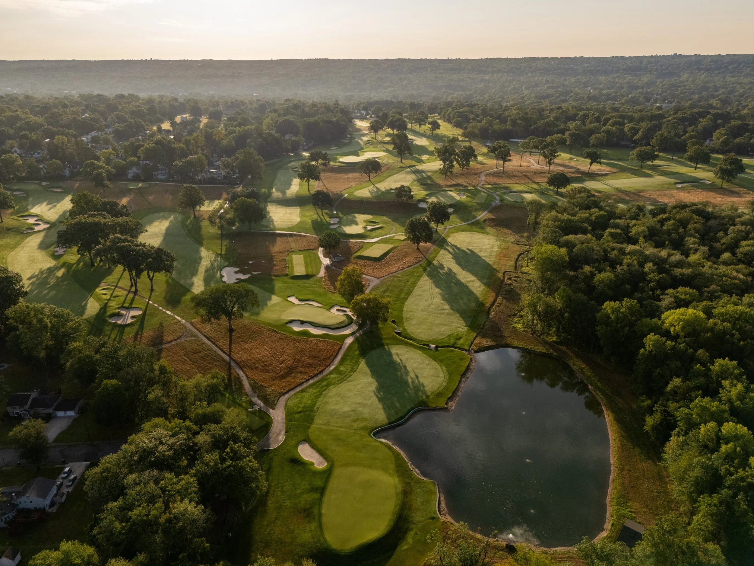An aerial view of a golf course with green fairways, sand traps, trees, and a large pond, with surrounding residential neighborhood and wooded areas.