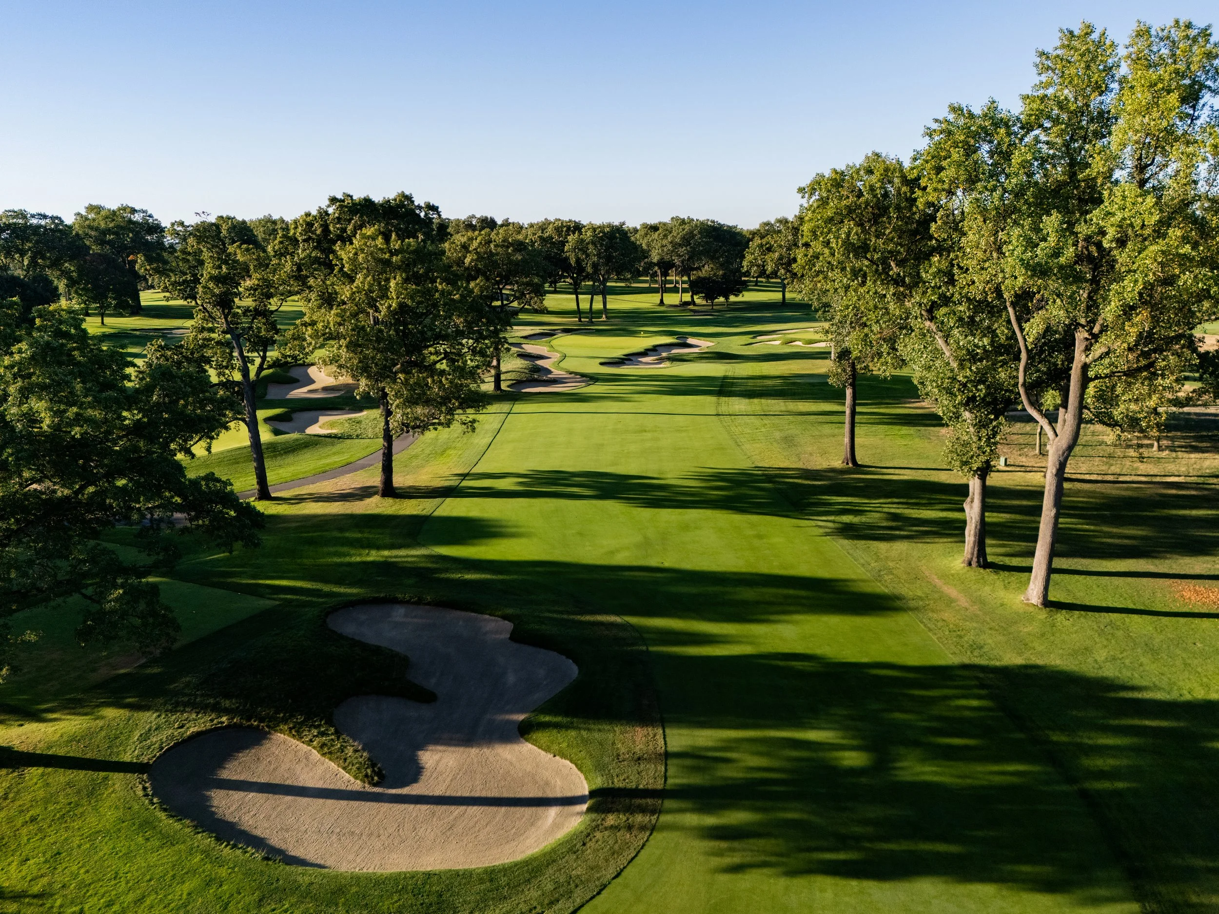 A golf course with green fairways, sand bunkers, and large trees under a clear blue sky.