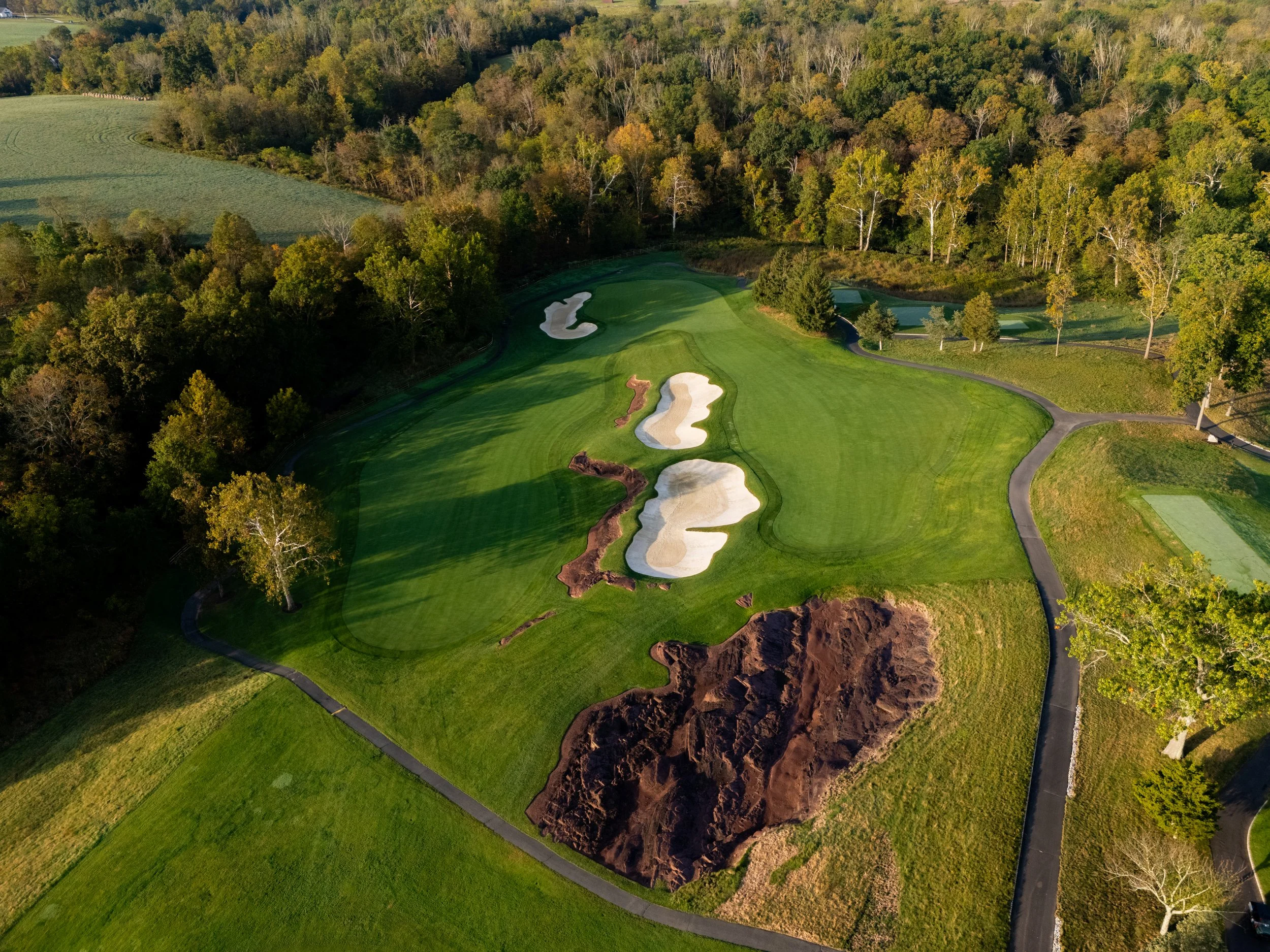An aerial view of a golf course, showing green fairways, sand traps, and a large area of dirt or construction site near the bottom.