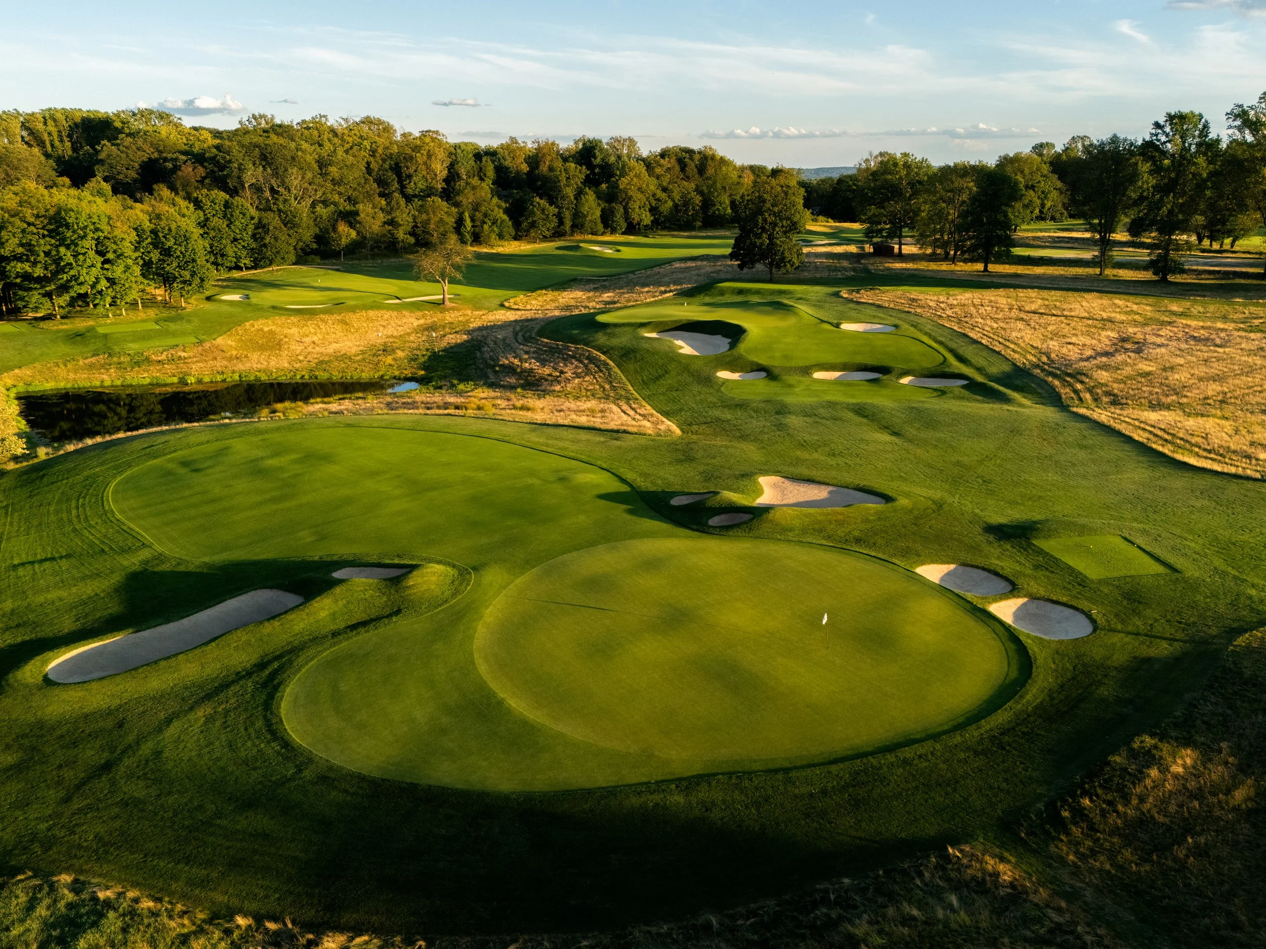 An aerial view of a golf course featuring green fairways, putting greens, and sand bunkers, surrounded by trees and open fields under a blue sky.