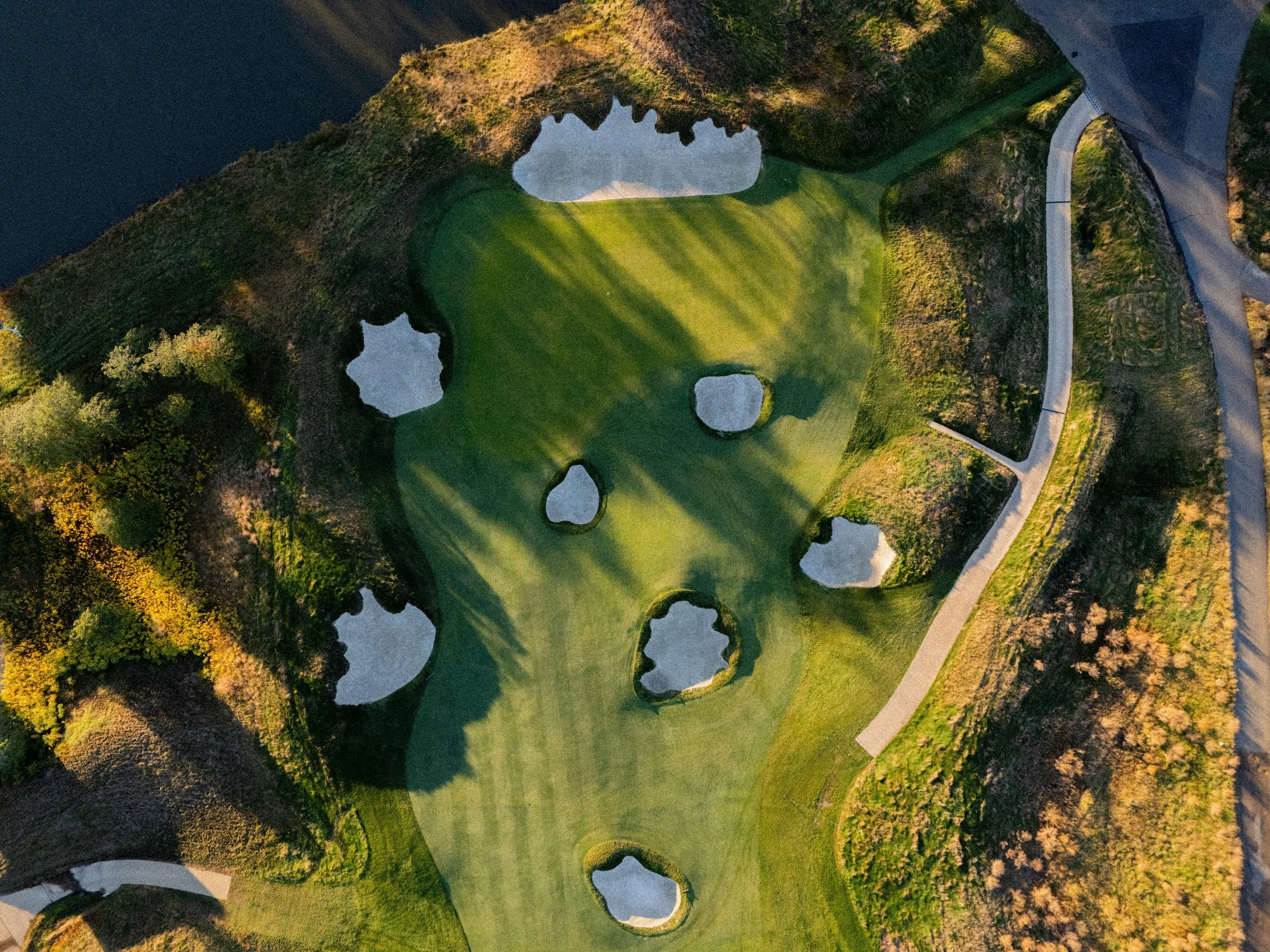 Aerial view of a golf course with multiple sand bunkers surrounding the green, located on a hillside with trees and walking paths.