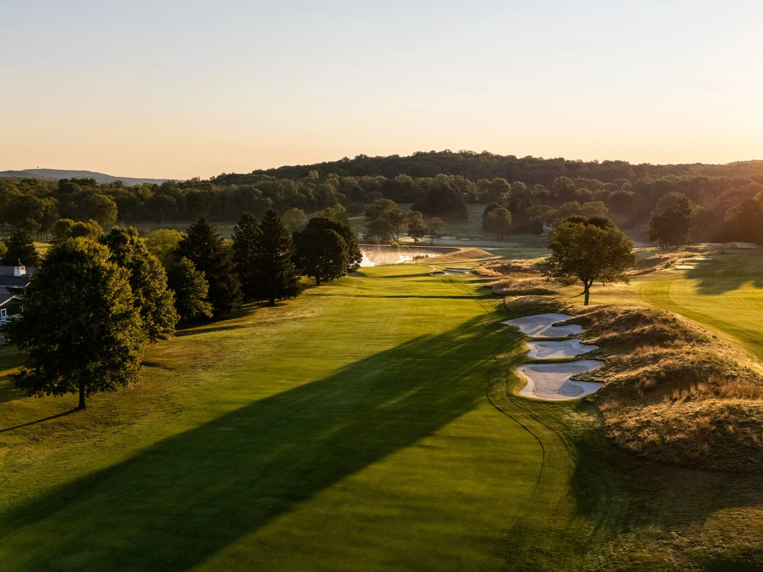 A golf course with green fairways, sand bunkers, and large trees under a clear blue sky.