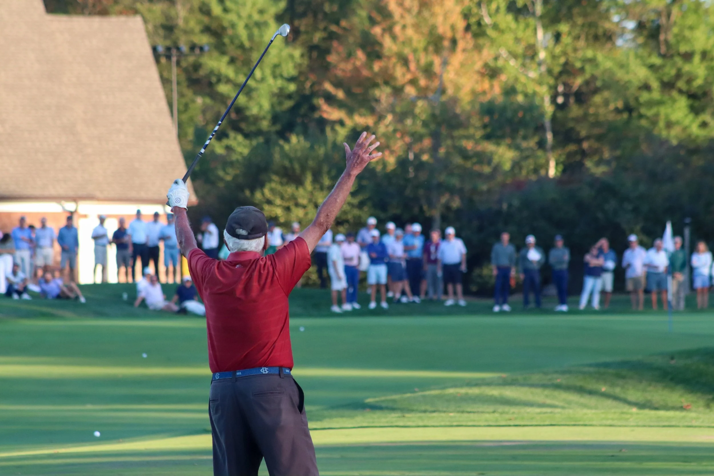 A golfer in a red shirt and black cap swings a golf club on a green golf course, with a crowd of spectators watching in the background.