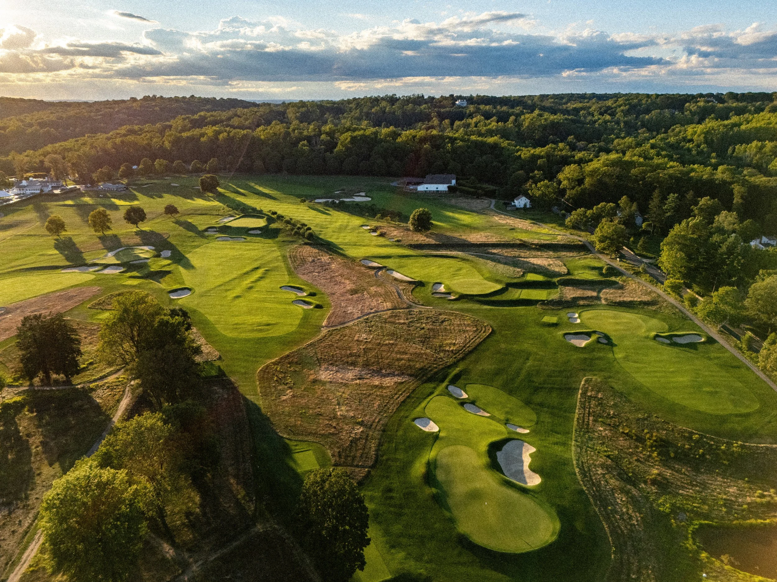 Aerial view of a golf course with green fairways, sand traps, and trees, surrounded by a wooded area under a partly cloudy sky during sunset.