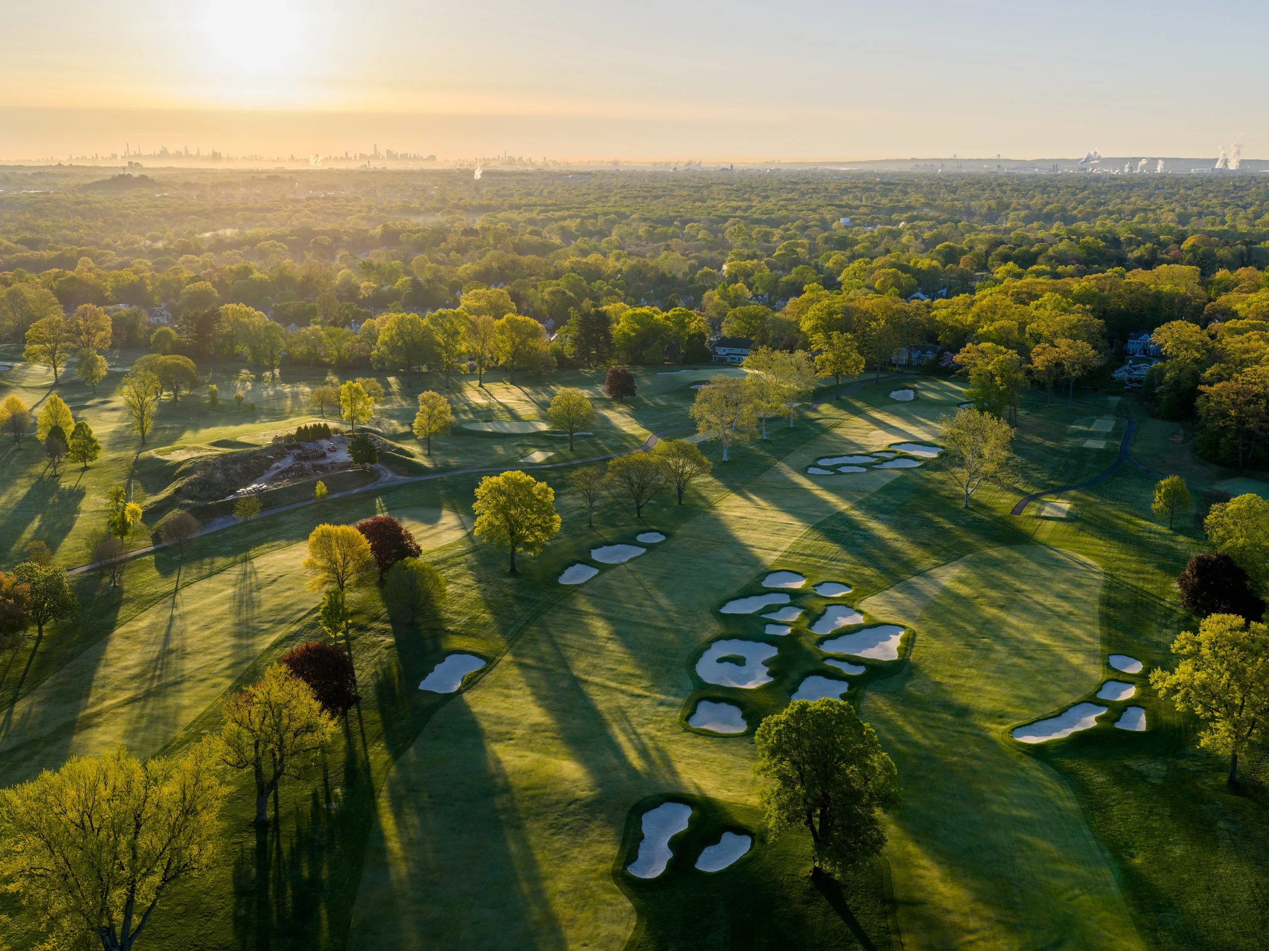 Aerial view of a golf course by a large body of water during sunset, with trees, pathways, and residential houses nearby.