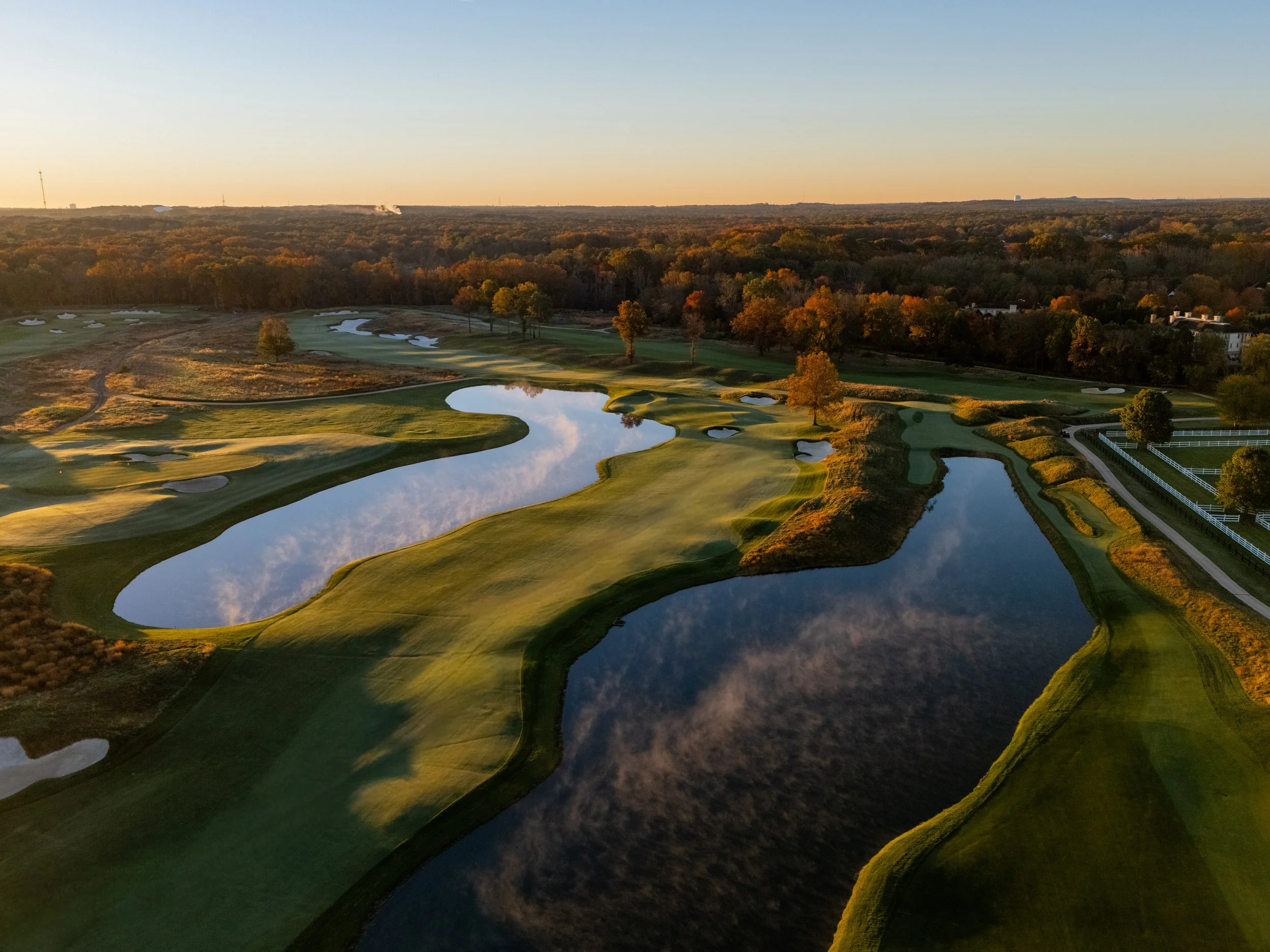 Aerial view of a golf course at sunset, featuring water hazards, sand bunkers, and green fairways surrounded by trees.