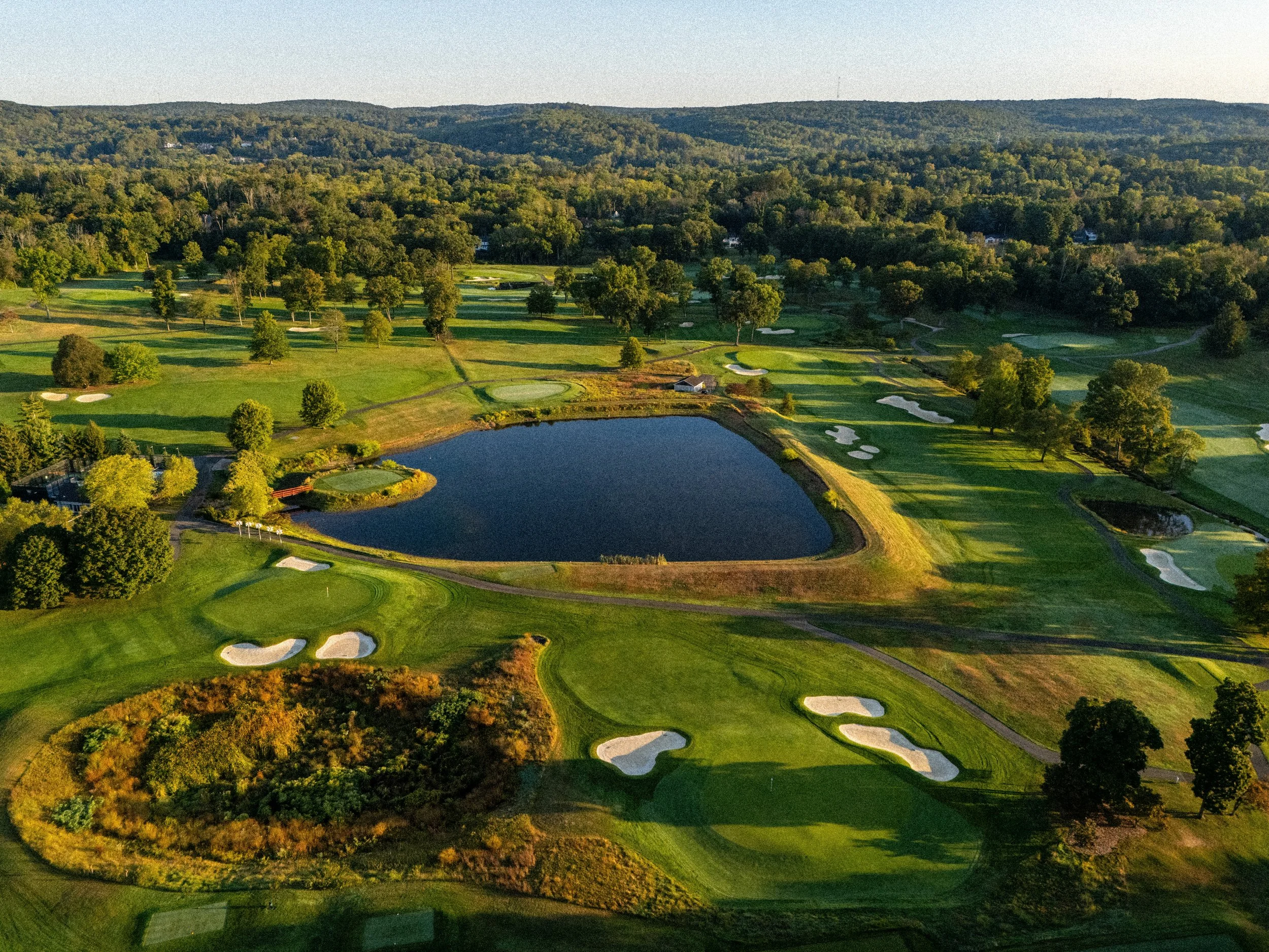 An aerial view of a golf course with multiple sand traps, water hazards, lush green fairways, and trees, set in a hilly landscape.