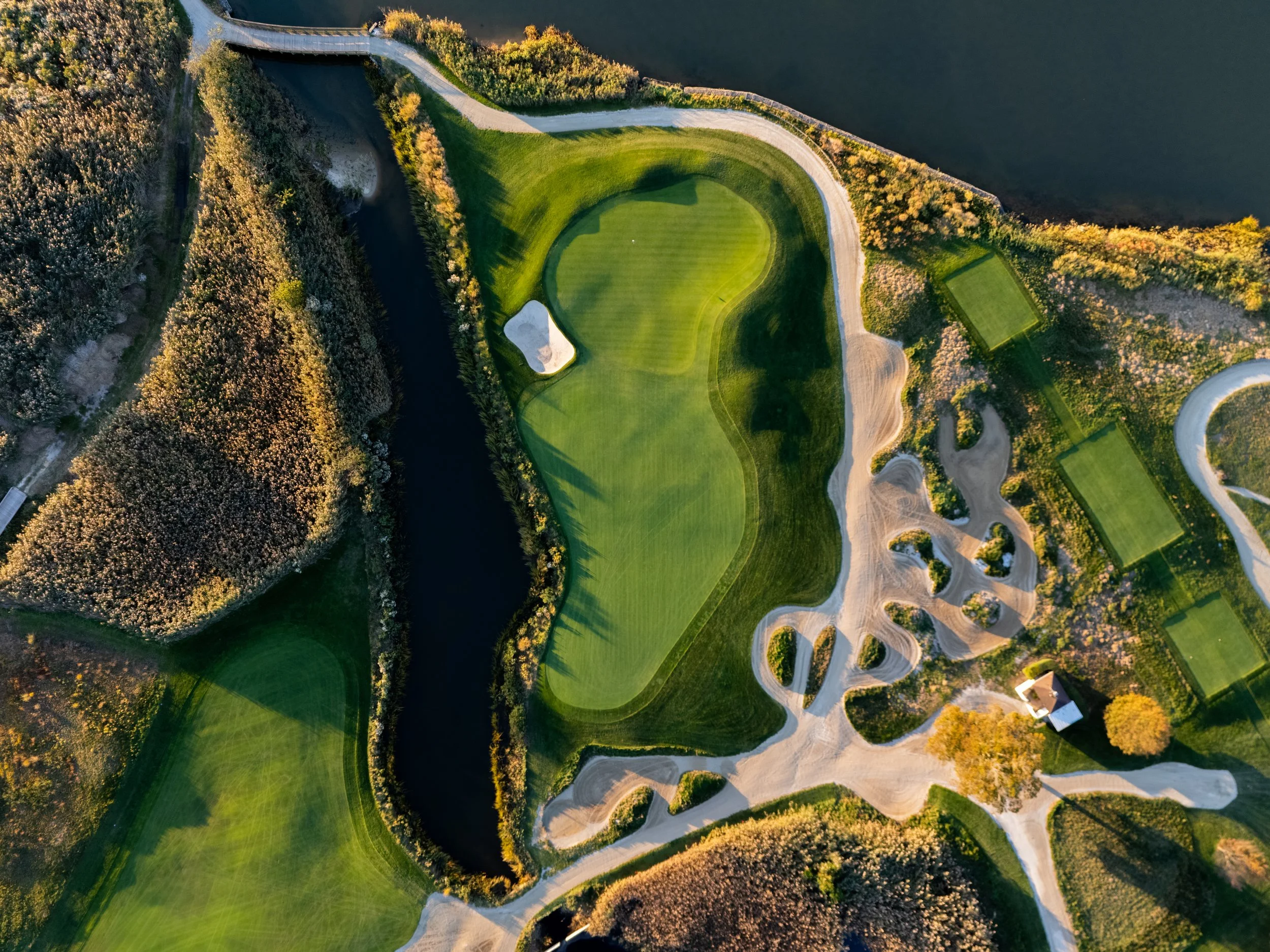Aerial view of a golf course with a green, sand bunkers, and water hazard, surrounded by trees and pathways.