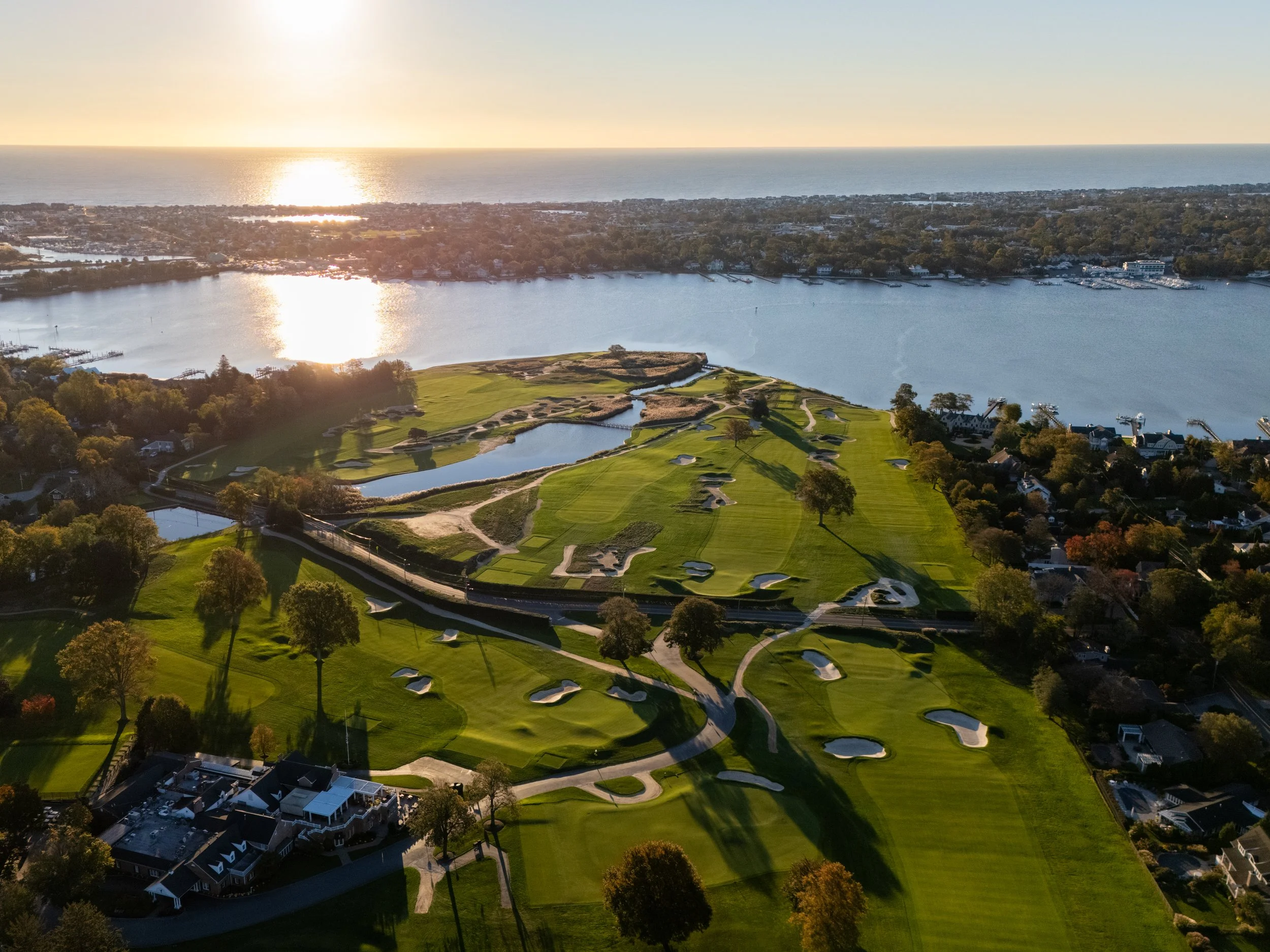 Aerial view of a golf course by a large body of water during sunset, with trees, pathways, and residential houses nearby.