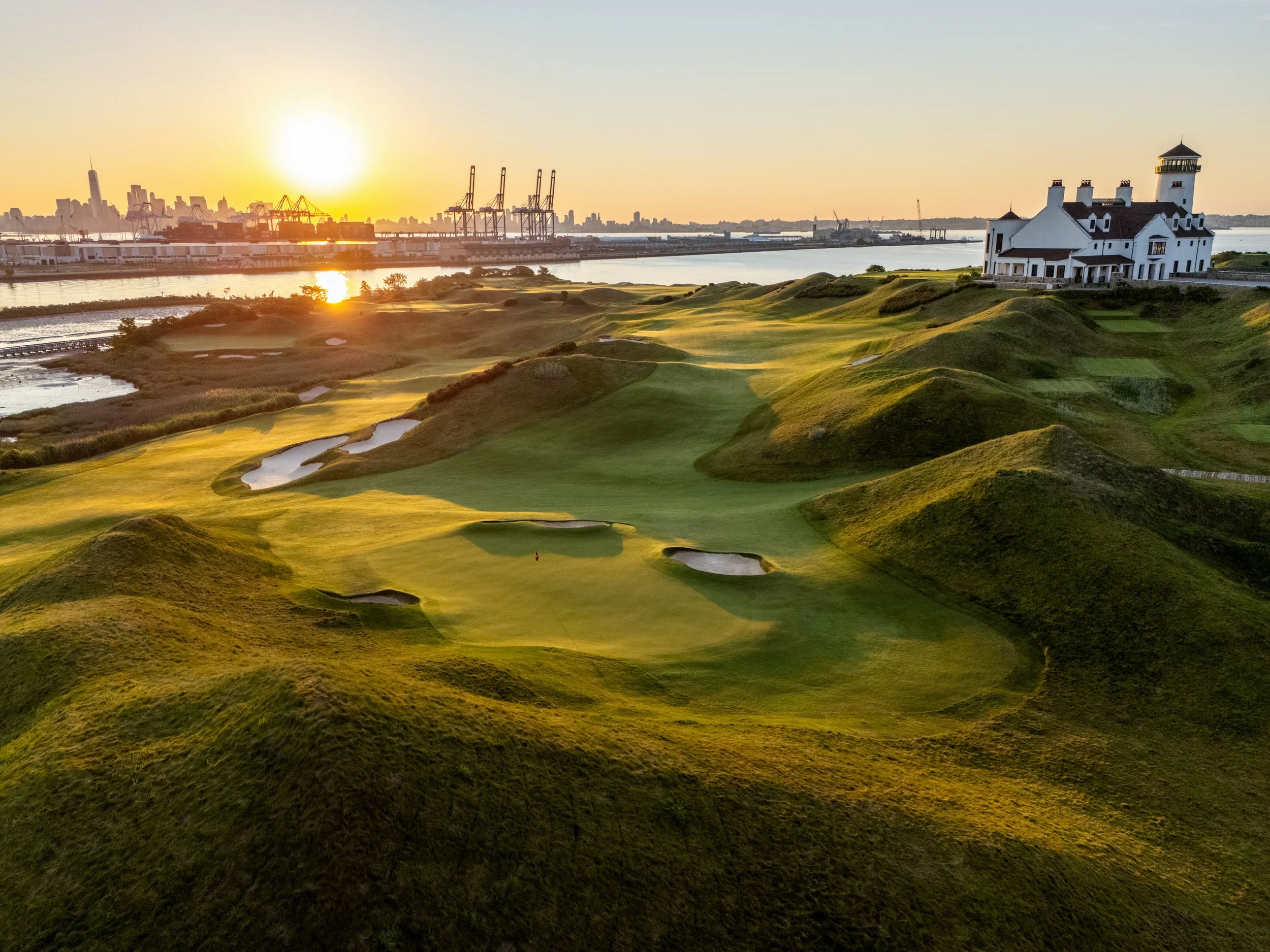 Sunset over a golf course with rolling green hills and sand traps near a body of water, with a white lighthouse-style building on the right and a city skyline with cranes in the background.