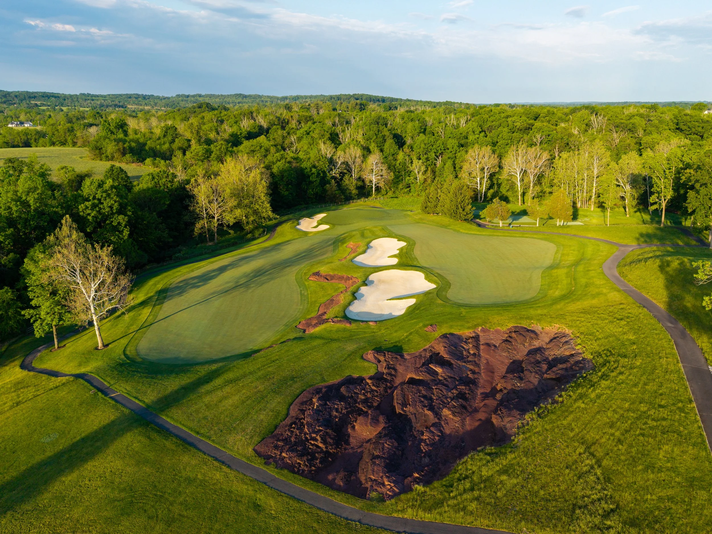An aerial view of a golf course, showing green fairways, sand traps, and a large area of dirt or construction site near the bottom.