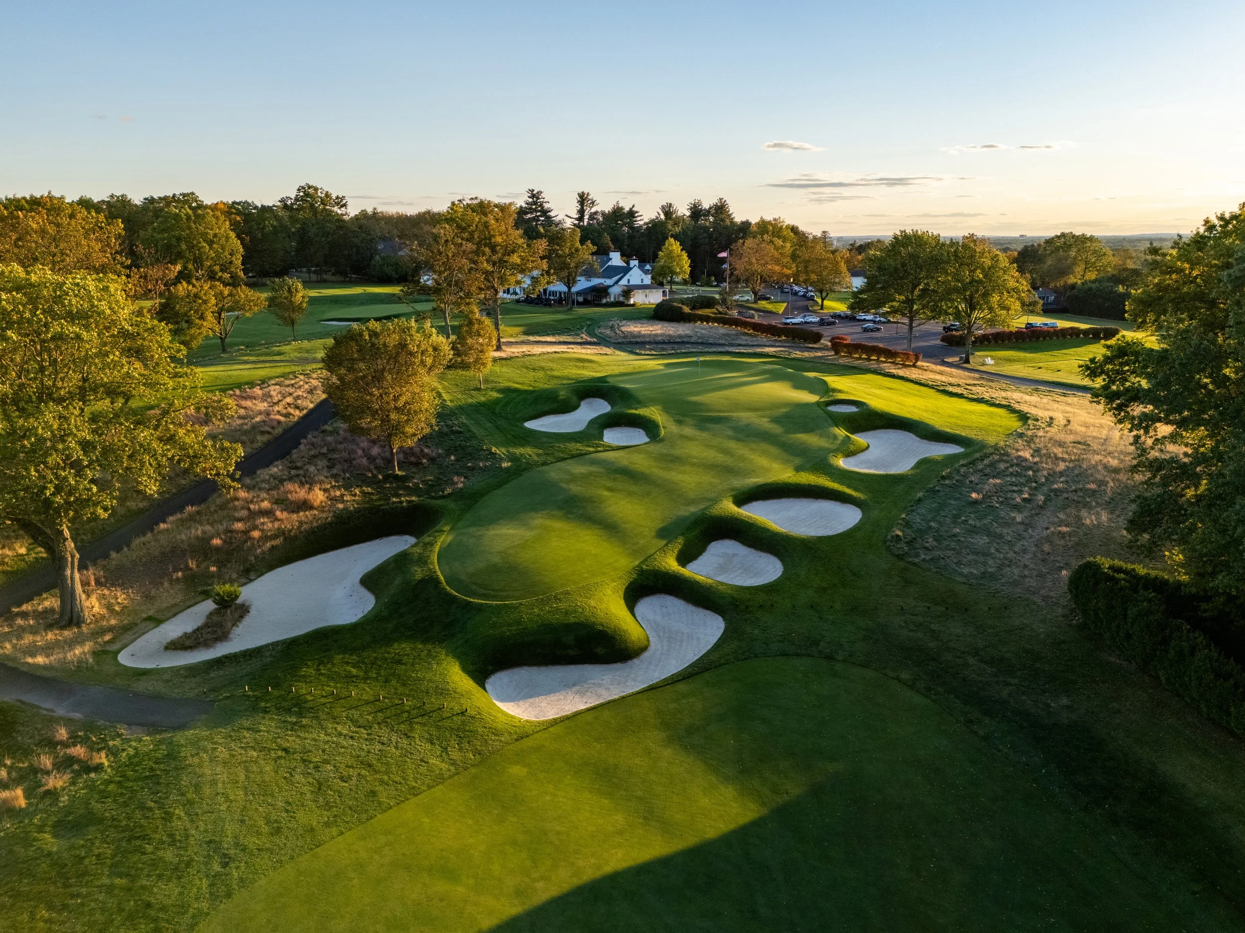 Aerial view of a golf course during sunset, with lush green grass, sand bunkers, trees, a clubhouse, and parking lot in the background.