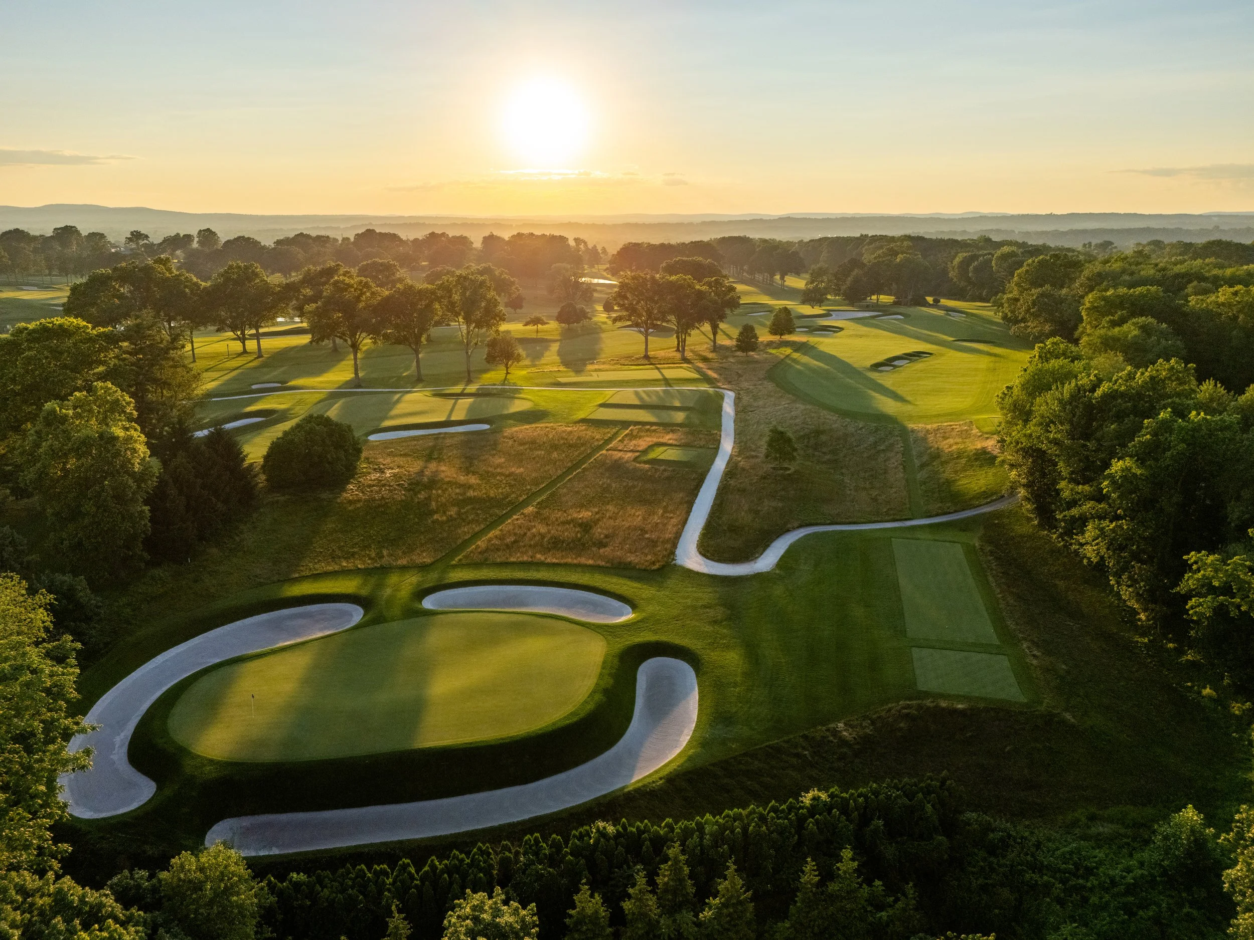 An aerial view of a golf course during sunset, featuring green putting greens, sand traps, and surrounded by trees.