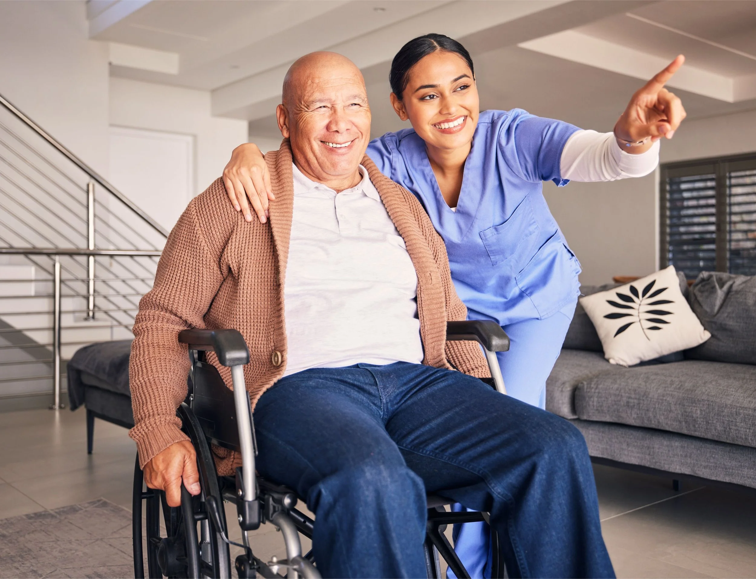 A smiling nurse in blue scrubs pointing at something while a happy older man in a wheelchair looks on inside a modern home.