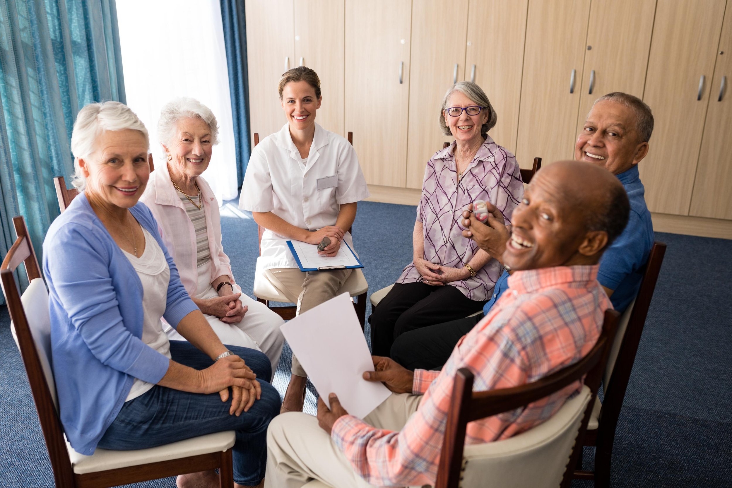 A diverse group of six elderly adults and a caregiver or medical professional smiling and sitting in a circle in a medical or care facility room.