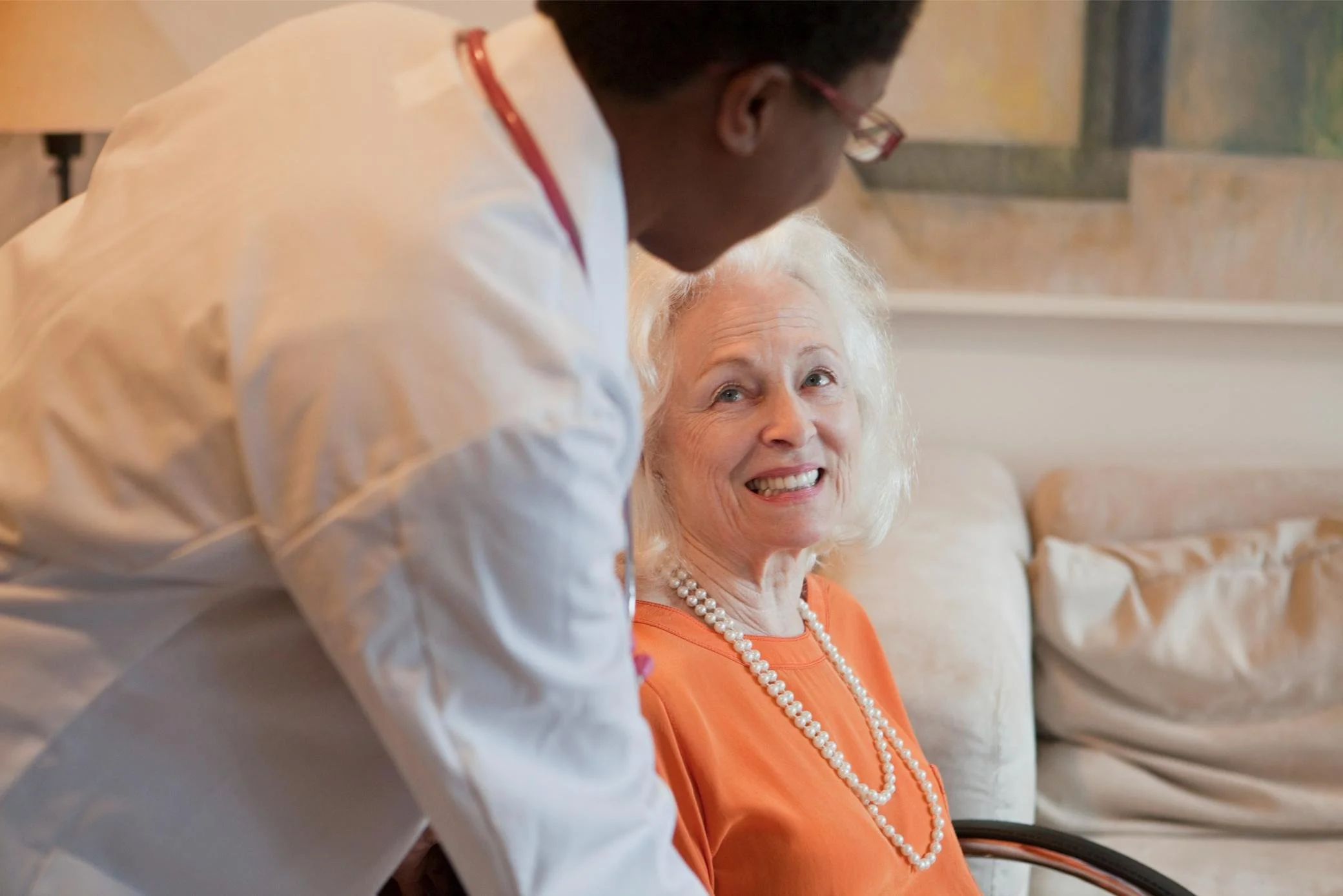 A healthcare worker in white uniform and red lanyard leaning towards an elderly woman with white hair, wearing an orange top and pearl necklace, who is sitting on a beige sofa and smiling.