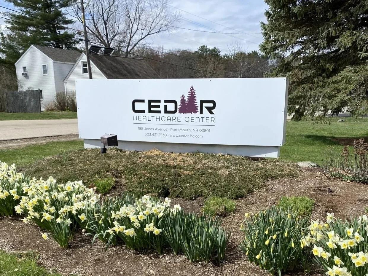 Sign for Cedar Healthcare Center with trees and flowers in front, residential houses in the background.