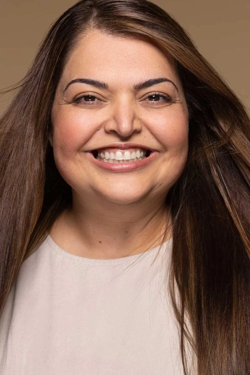 A woman with long brown hair smiling at the camera, wearing a light-colored top.