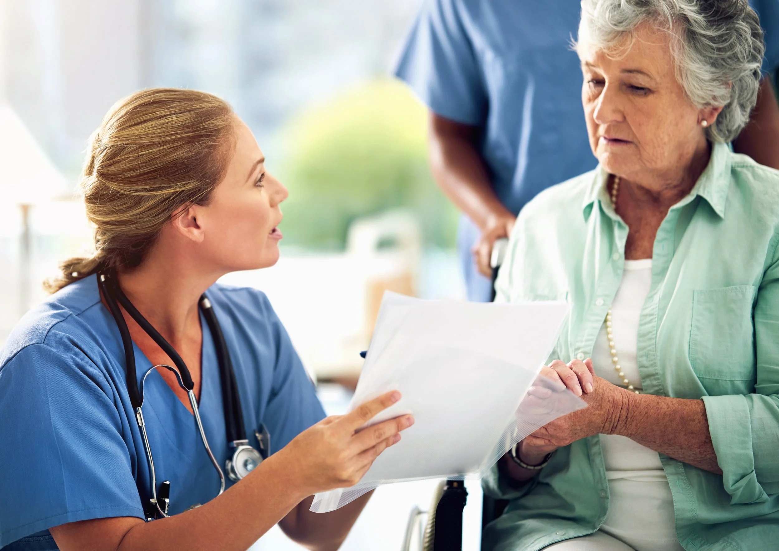 A nurse in blue scrubs with a stethoscope around her neck holding a medical chart, talking to an elderly woman with gray hair and a pearl necklace, who is in a wheelchair. A healthcare worker in blue scrubs stands behind them in a brightly lit facility.