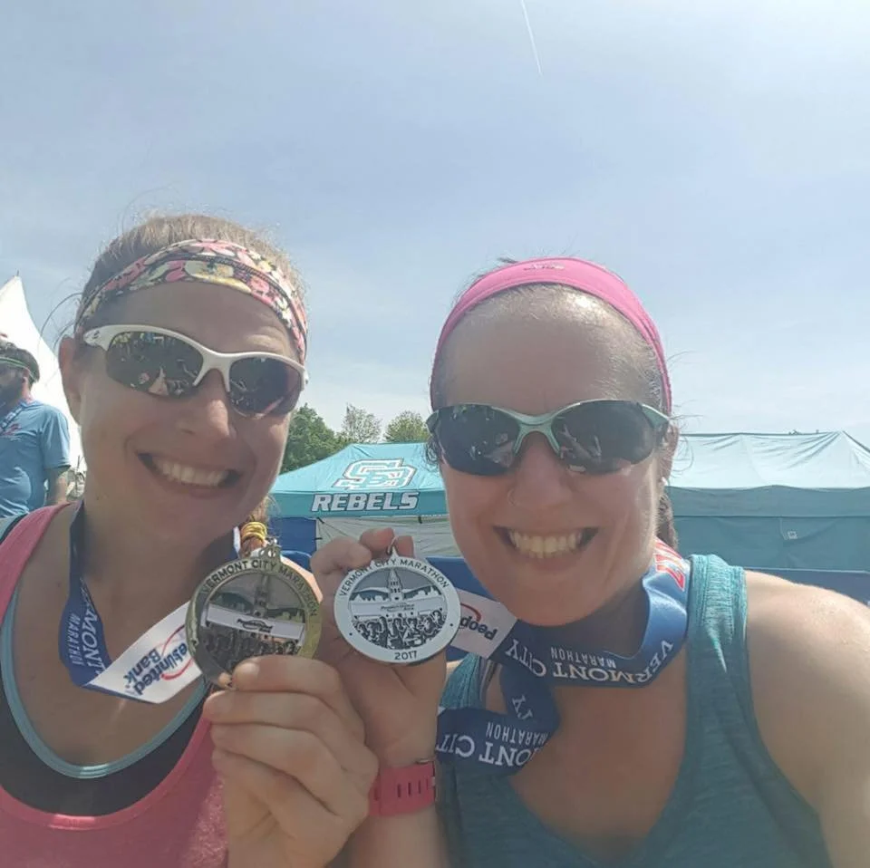Two women at a race event, holding marathon medals, smiling, wearing sports gear, sunglasses, and headbands, with race tents in the background.