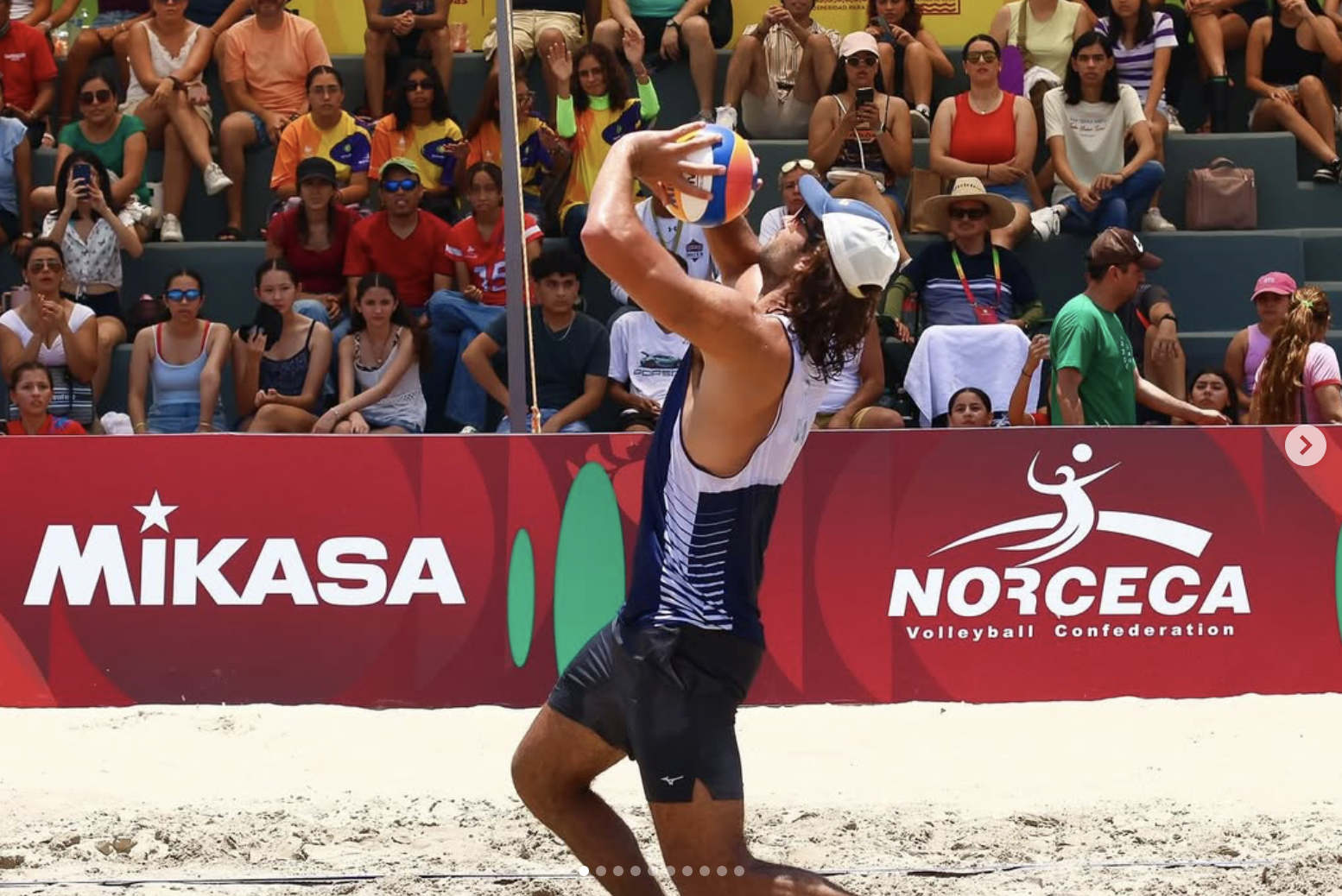 A beach volleyball player in a white tank top and black shorts prepares to hit the volleyball during a match. The background shows a crowd of spectators seated and watching, with some taking photos in front of a red advertising banner for Mikasa and Norceca Volleyball Confederation.