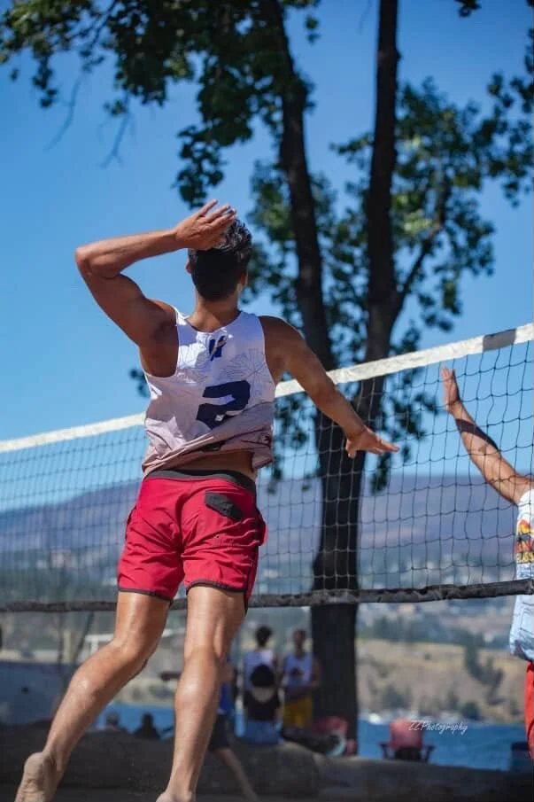 A male volleyball player is jumping to spike the ball at the net during a match outdoors, with trees and a blue sky in the background.